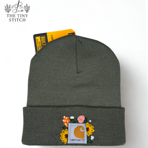 A dark green Carhartt beanie with floral embroidery on the front and a Carhartt logo patch, photographed on a white background with a tag from The Tiny Stitch in the top left corner.