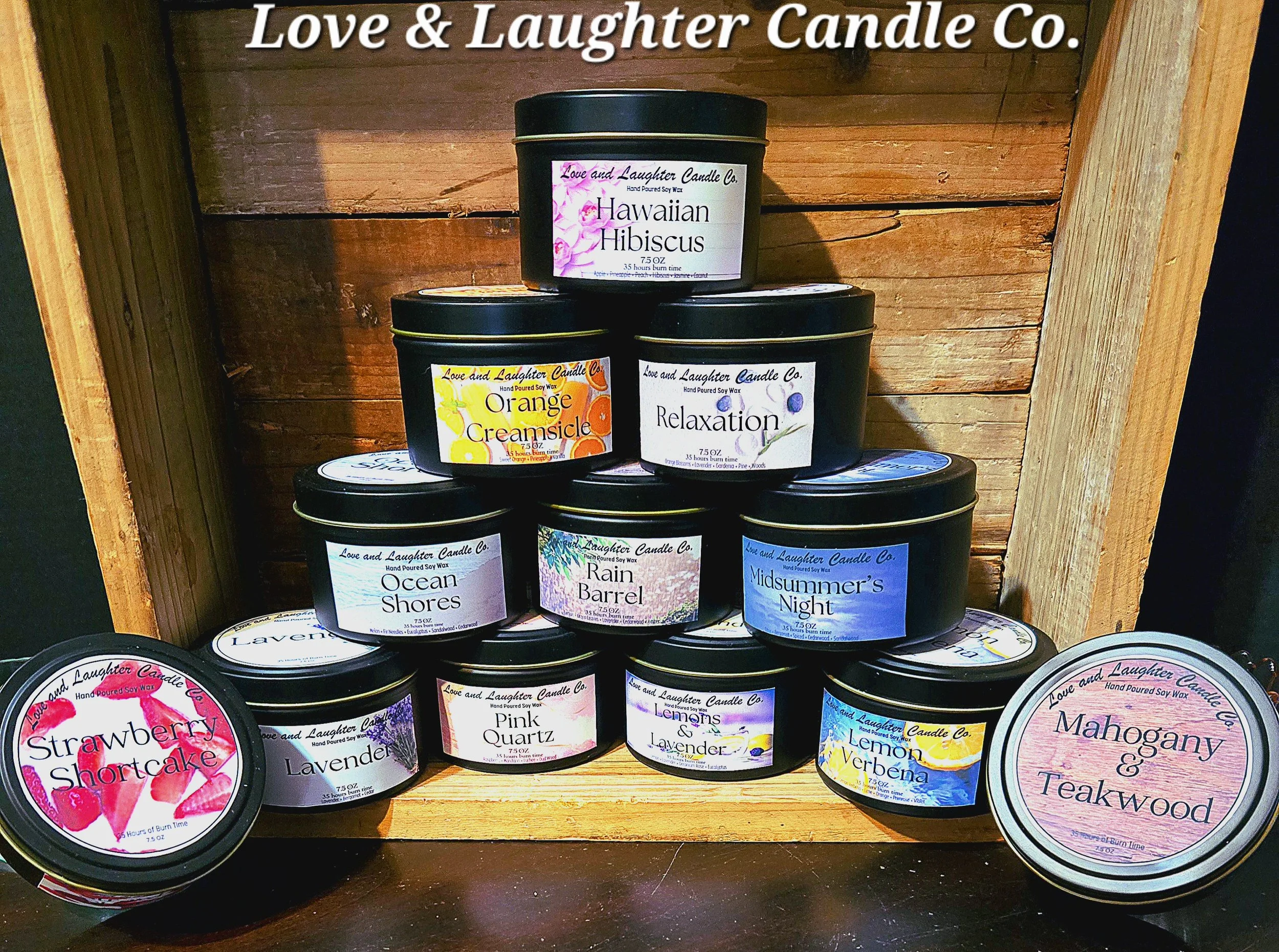 A display of scented candles in black containers with colorful labels, arranged on a wooden shelf. The candles are from Love & Laughter Candle Co. with scents including Hawaiian Hibiscus, Orange Creamsicle, Relaxation, Ocean Shores, Rain Barrel, Mids
