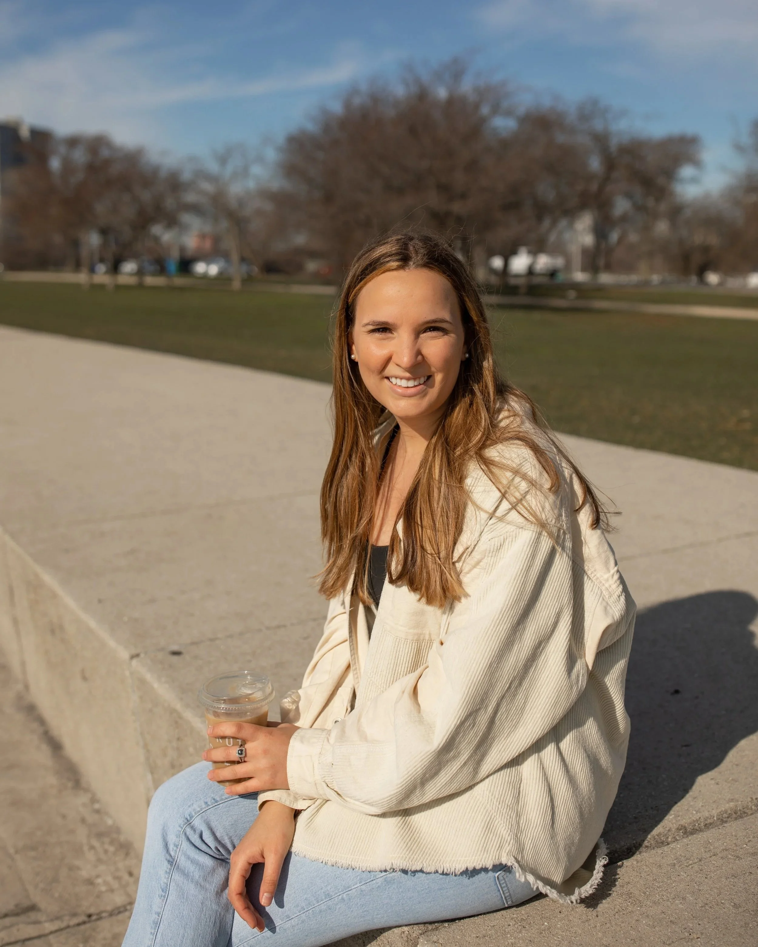A young woman with long brown hair, wearing a cream-colored jacket and light blue jeans, sitting on a concrete ledge in a park on a sunny day, holding a transparent cup of iced coffee.
