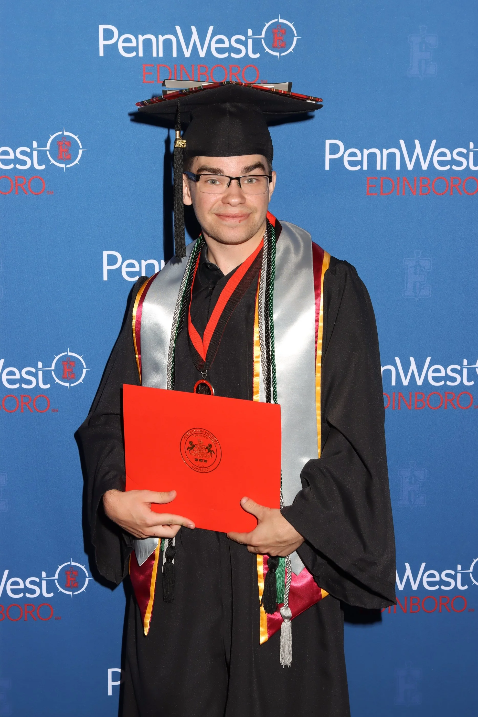 A young man in cap and gown holding a red diploma folder during graduation ceremony at PennWest Edinboro, standing in front of a blue graduation backdrop with PennWest Edinboro logos.