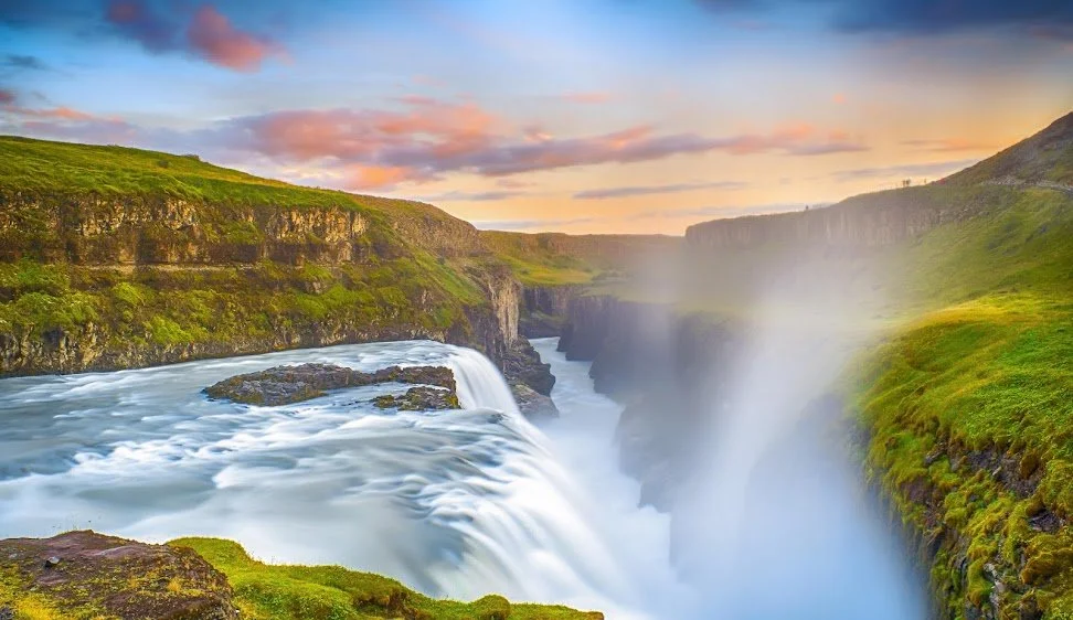 Waterfall flowing into a canyon with green grass and cliffs, under a colorful sky.