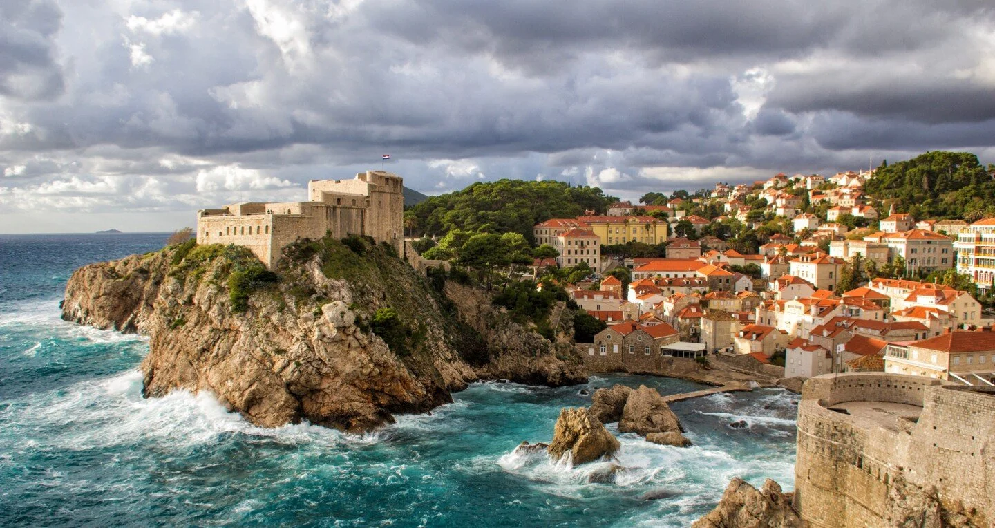 A coastal town with historical fortress on a rocky cliff overlooking the ocean, with tightly packed buildings with red-tiled roofs and a cloudy sky overhead.