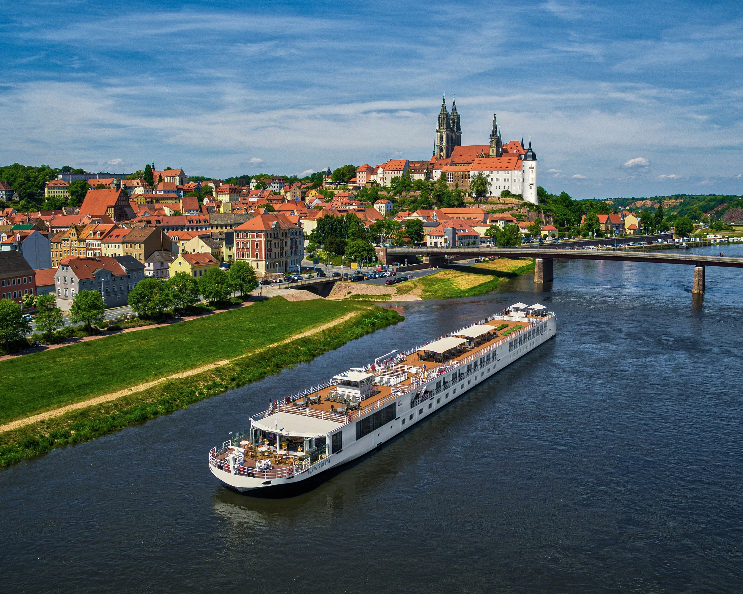 A river with a large white boat sailing on it, flowing through a town with colorful buildings and a large church on a hill in the background under a blue sky with scattered clouds.