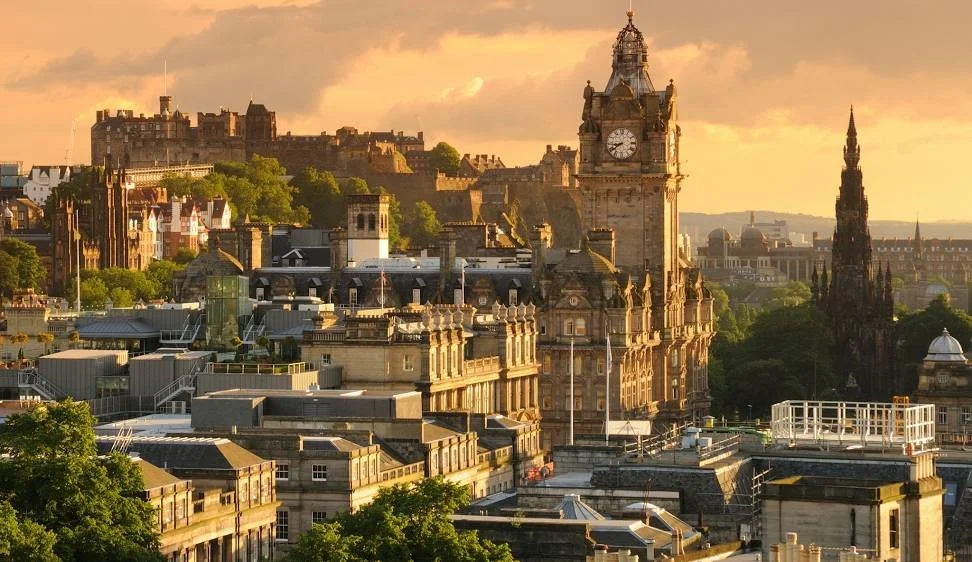 Sunset view of Edinburgh cityscape featuring the clock tower of the Palace of Holyroodhouse, historic buildings, and green hills in the background.