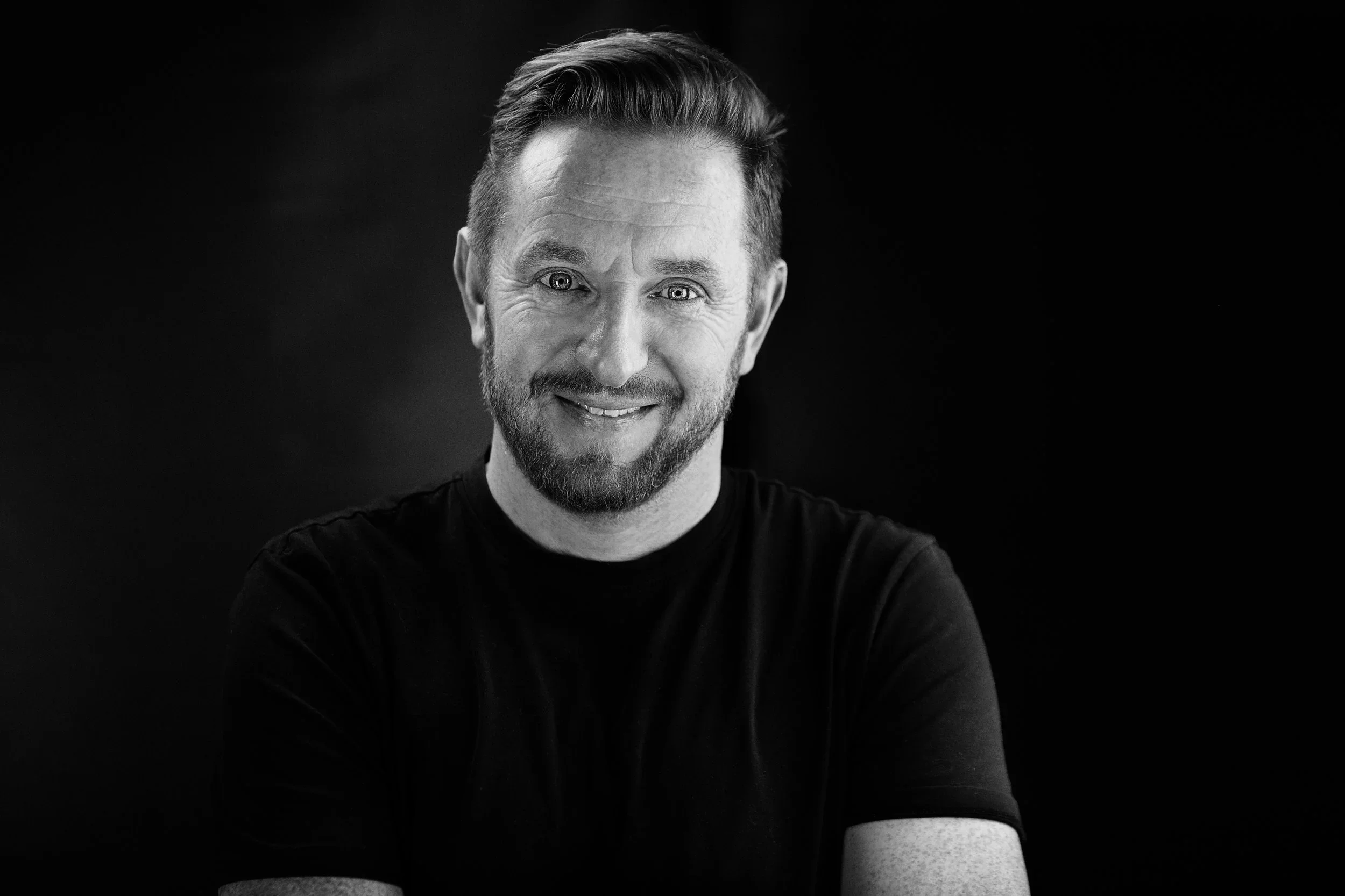 Black and white portrait of a smiling man with short hair and a beard, wearing a black shirt, against a dark background.
