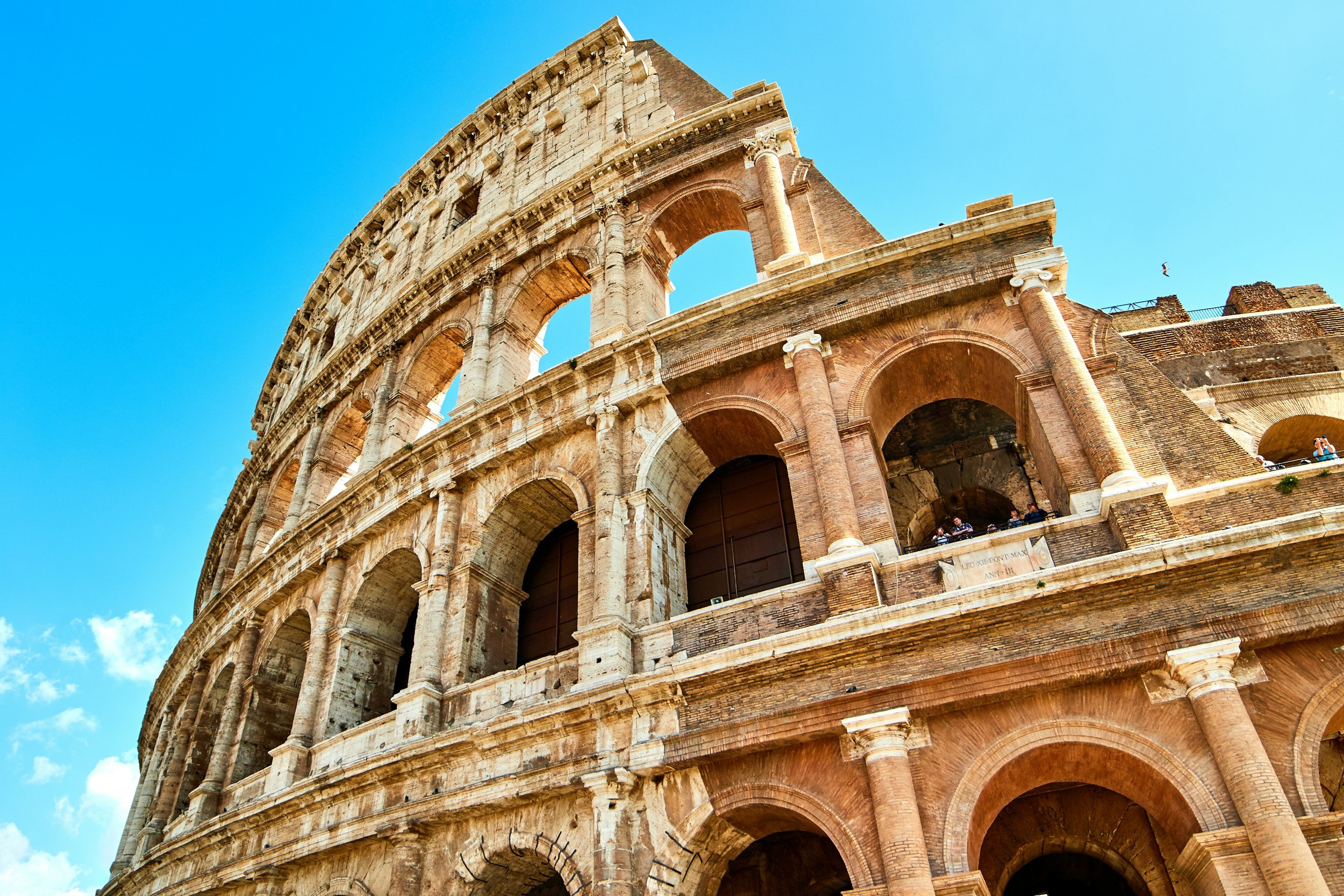 Close-up of the Roman Colosseum showing its arches and ancient brick and stone structure against a bright blue sky.
