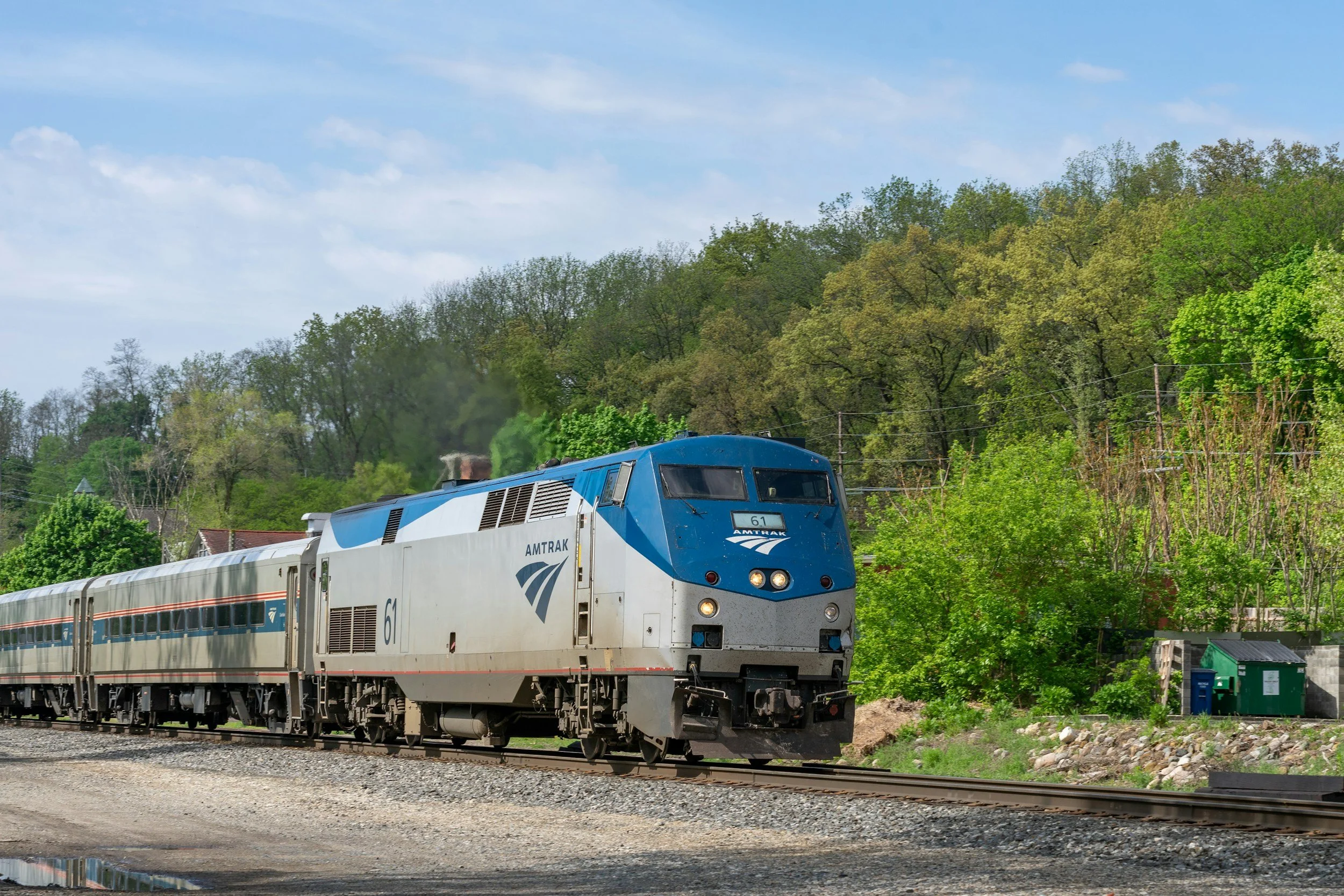 A blue and white Amtrak passenger train moving along railway tracks through a rural area with green trees and hills in the background.