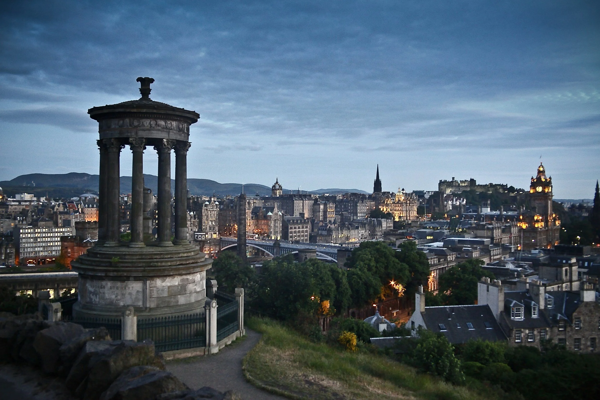 A cityscape of Edinburgh at dusk with historic buildings, a monument with columns in the foreground, and a cloudy sky.