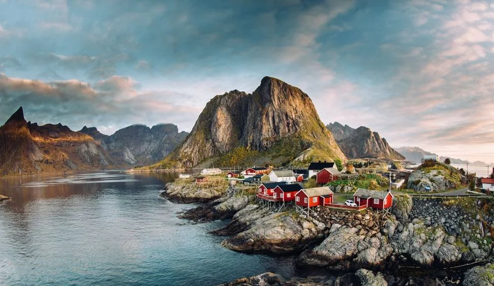 Coastal village with red houses on rocky shoreline surrounded by mountains and water under a partly cloudy sky.