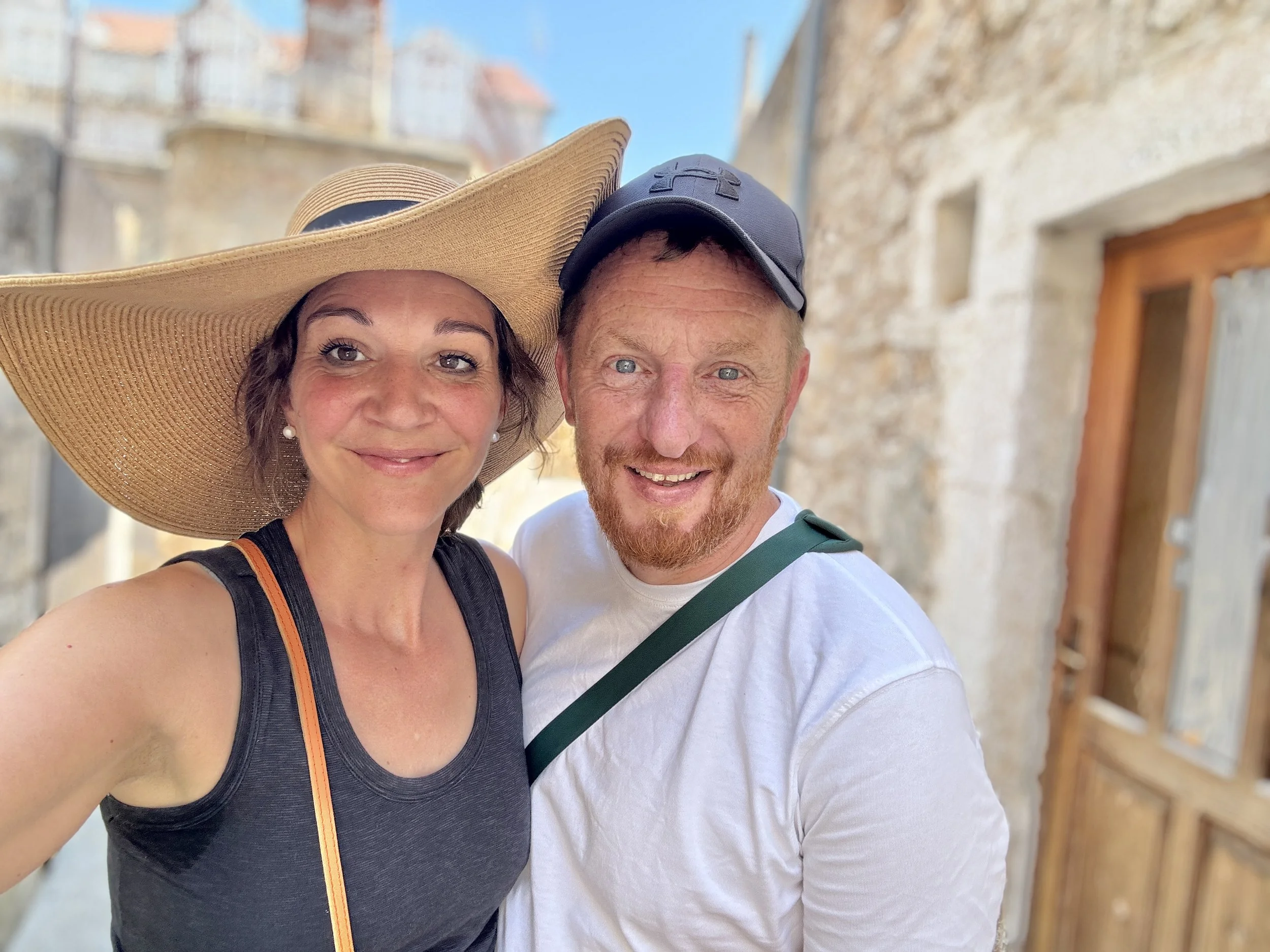 A woman and a man smiling for a selfie outdoors. The woman is wearing a large wide-brimmed hat and a sleeveless black top. The man is wearing a black cap, white shirt, and has a green strap across his chest. They are standing in front of an old stone building with a wooden door, under a clear blue sky.