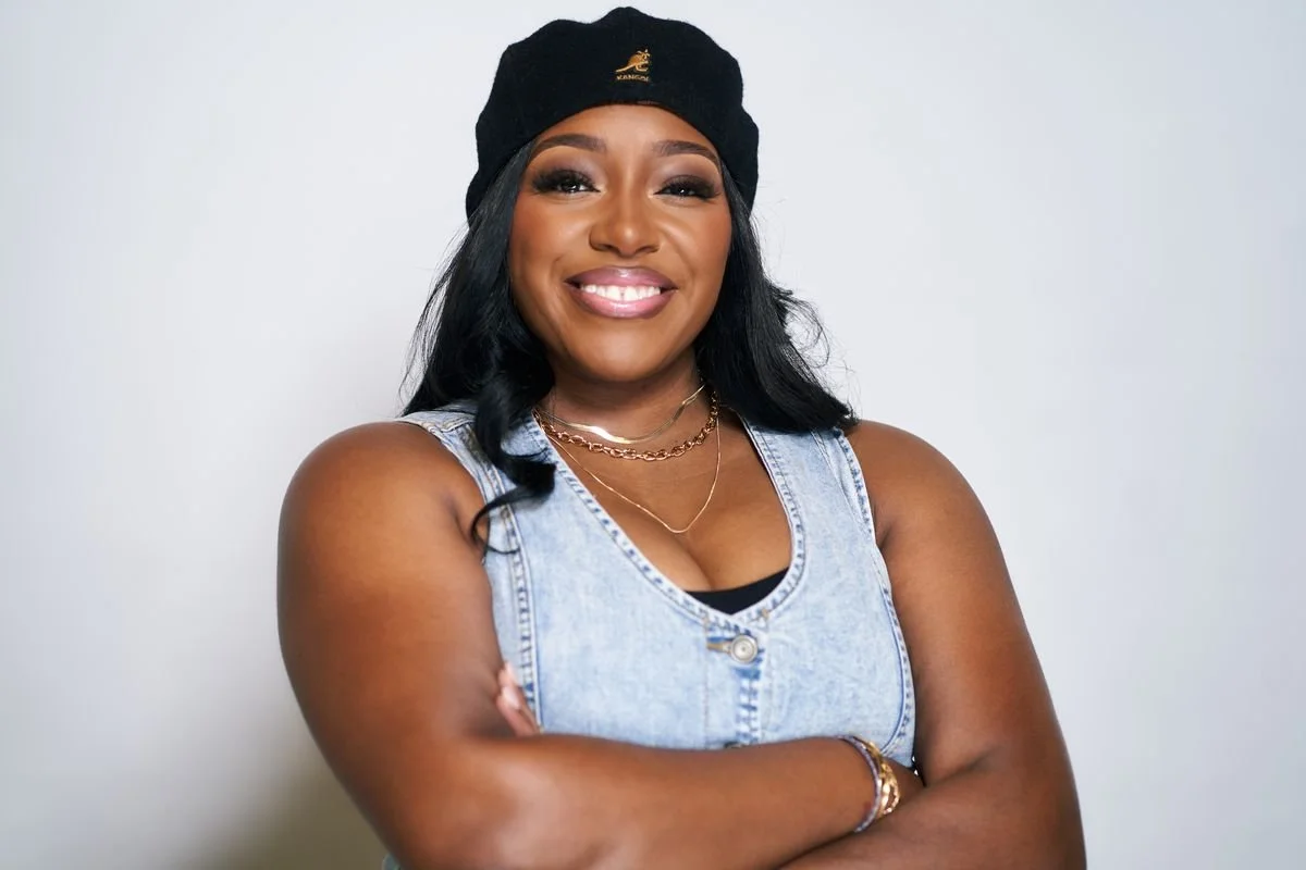 A woman with long black hair smiling, wearing a black beanie, denim sleeveless top, and gold jewelry, standing against a plain white background.