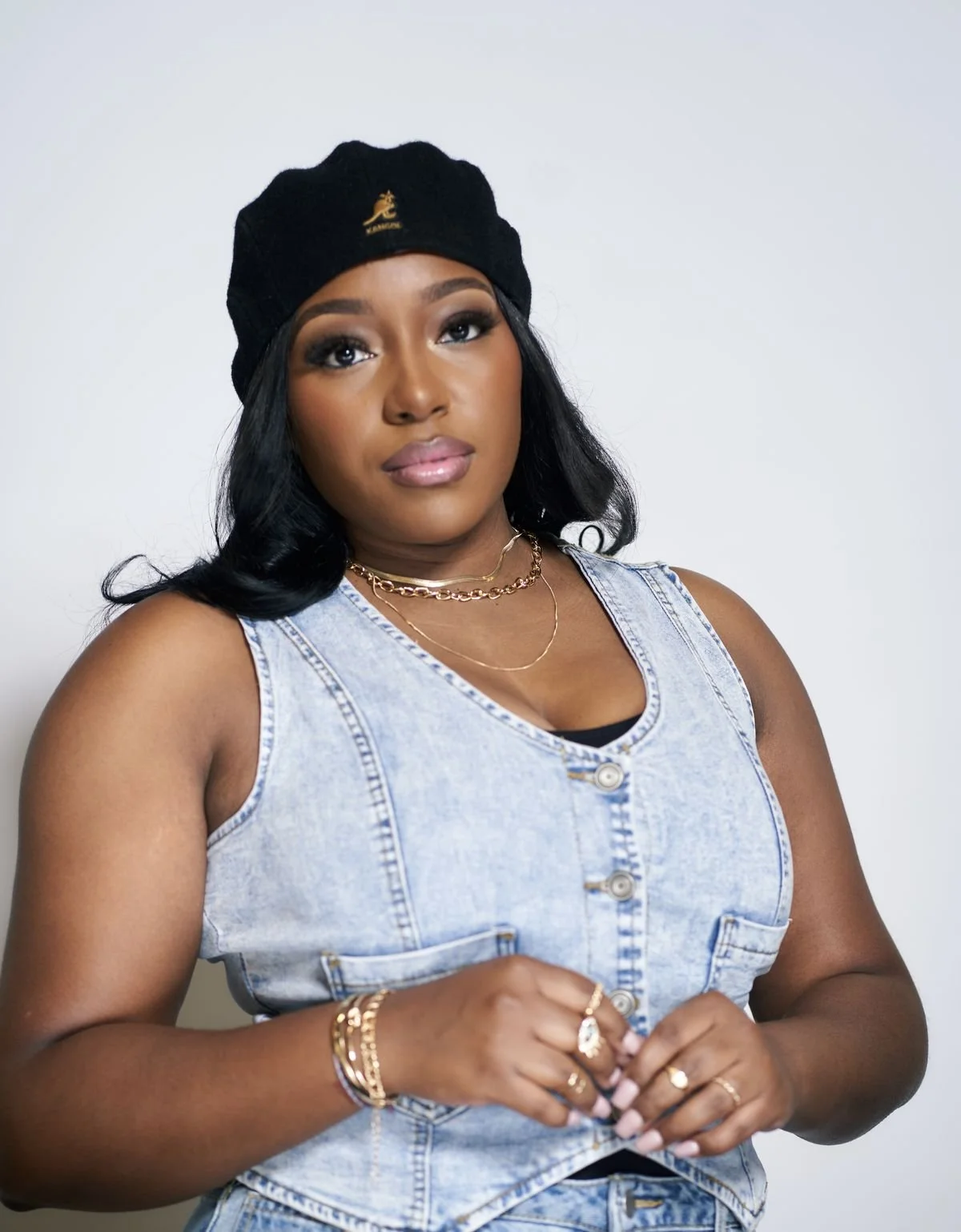 A woman with dark wavy hair wearing a black beret, layered gold necklaces, and gold jewelry on her hands, dressed in a sleeveless denim vest, standing against a white background.