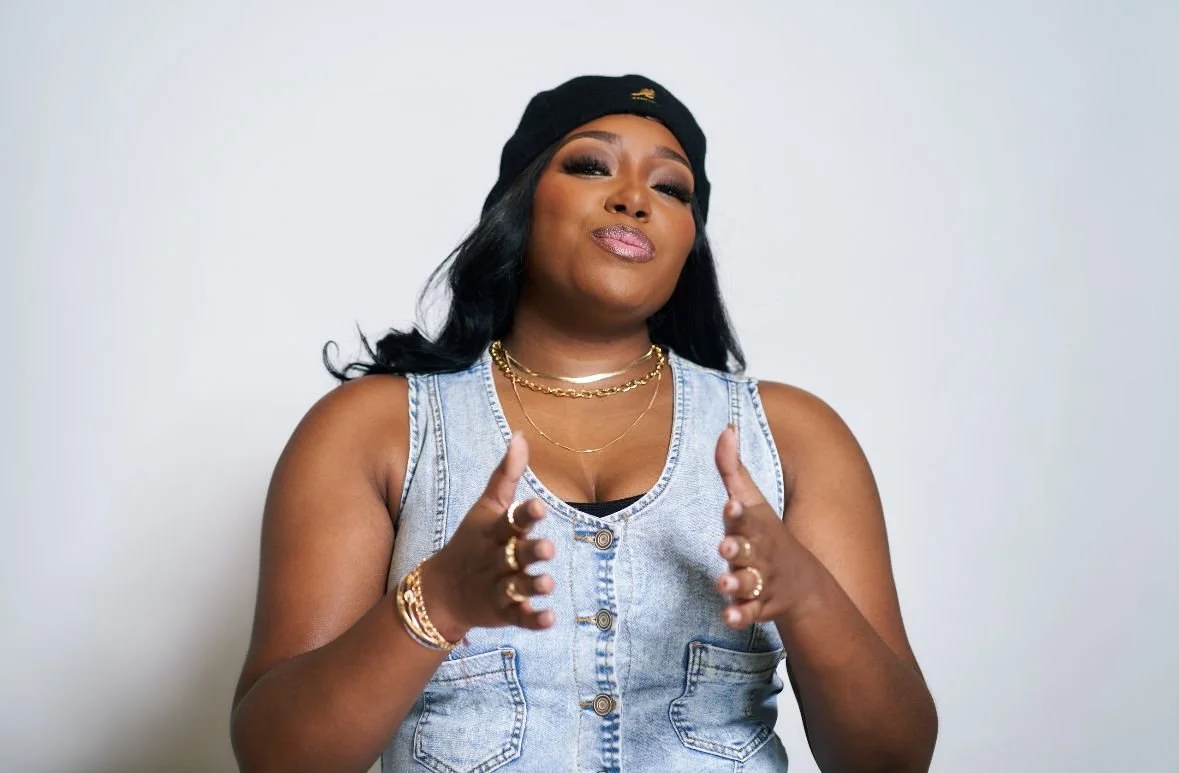 Woman with black hair wearing a black beanie, denim vest, and layered gold necklaces, gesturing with her hands against a plain white background.