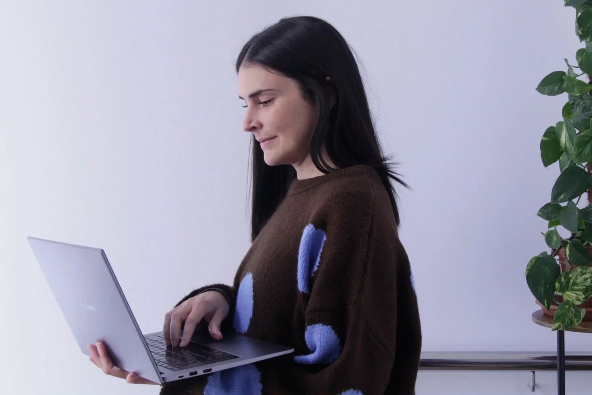 A woman with black hair wearing a brown sweater with blue patterns holding a silver laptop.