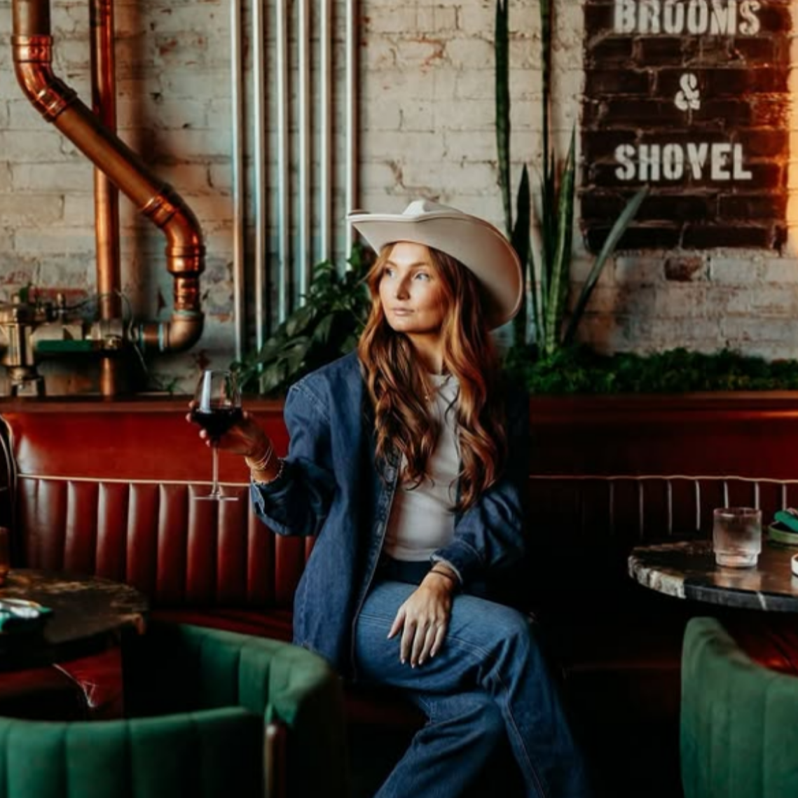 A woman with long red hair, wearing a large beige cowboy hat and denim outfit, sitting in a rustic bar or cafe, holding a glass of red wine, behind her a brick wall with industrial pipes and a sign that reads 'Brooms & Shovel'. she is looking to her 