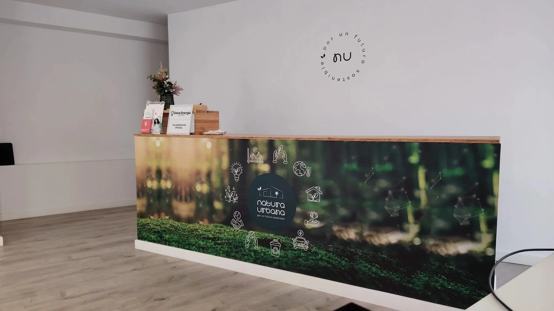 Reception desk with green nature themed graphic and icons, with a white wall that has a circular logo or sign with the text "por un futuro" and "sostenible".