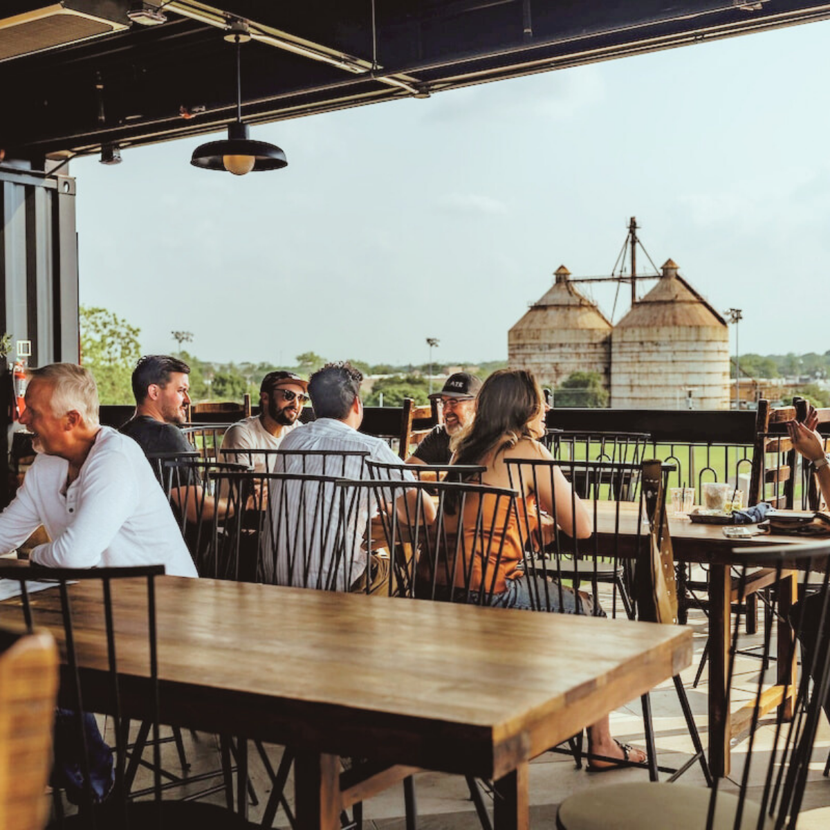 People sitting at a balcony dining area, enjoying conversations with a view of green fields and large barn structures in the background.