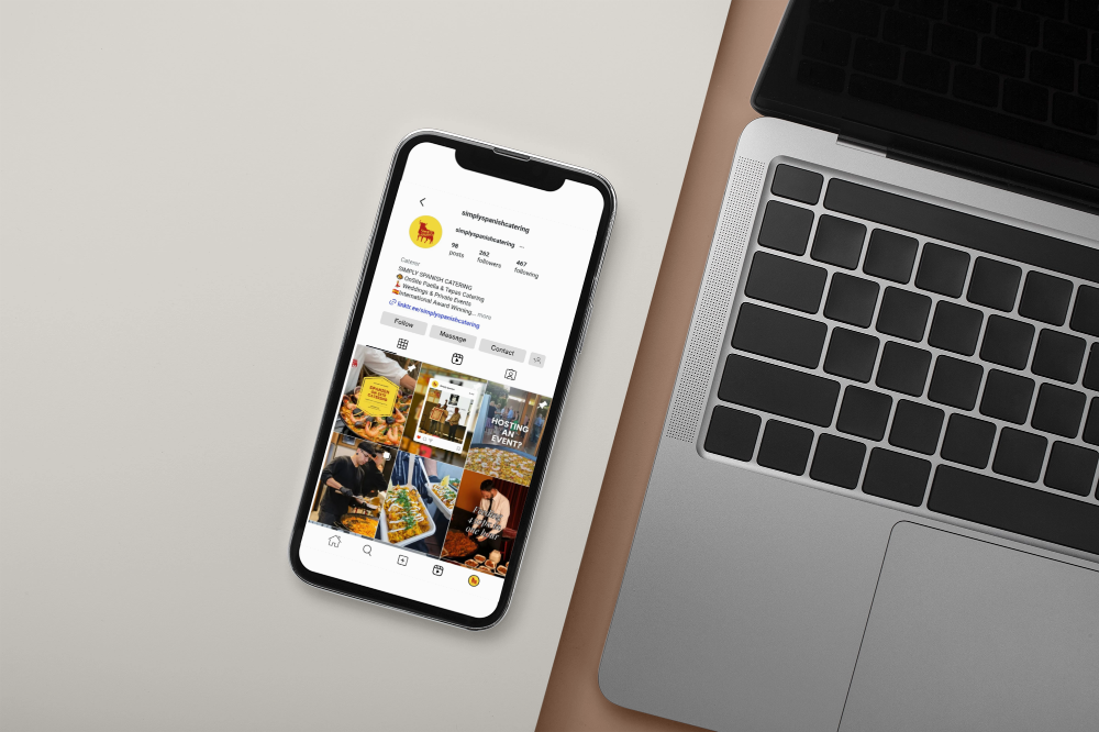 Smartphone displaying an Instagram profile for a catering business, with photos of food, event setups, and staff, placed next to a silver laptop on a beige surface.