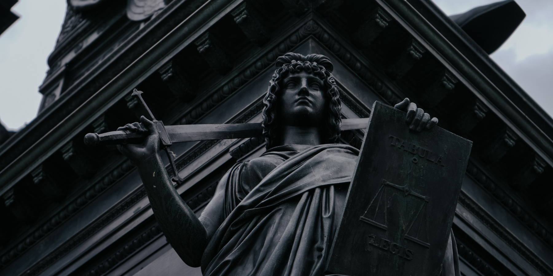Close-up of a statue of a woman holding a sword and a shield, with the shield inscribed with the word 'TABULA' and an image of scales, set against a building with ornate details.