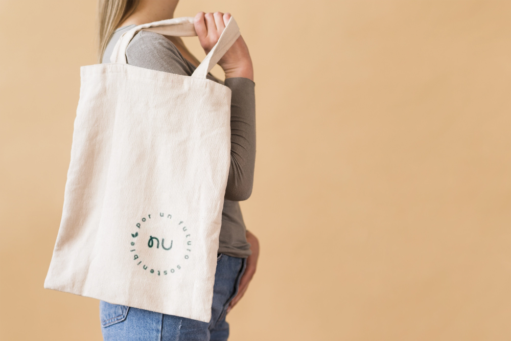 Person holding a beige tote bag over shoulder, wearing a gray shirt and jeans, against a neutral background.