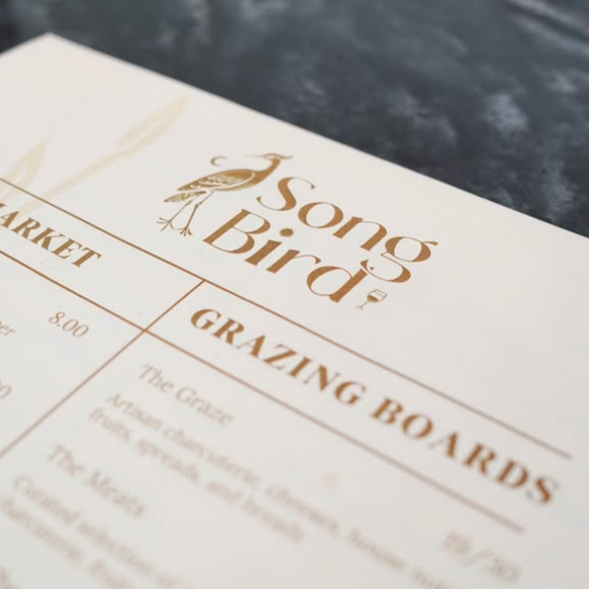 Close-up of a white menu with gold text for S.O.S Bird featuring a peacock logo, listing a grazing board at $8.00 and other menu items.
