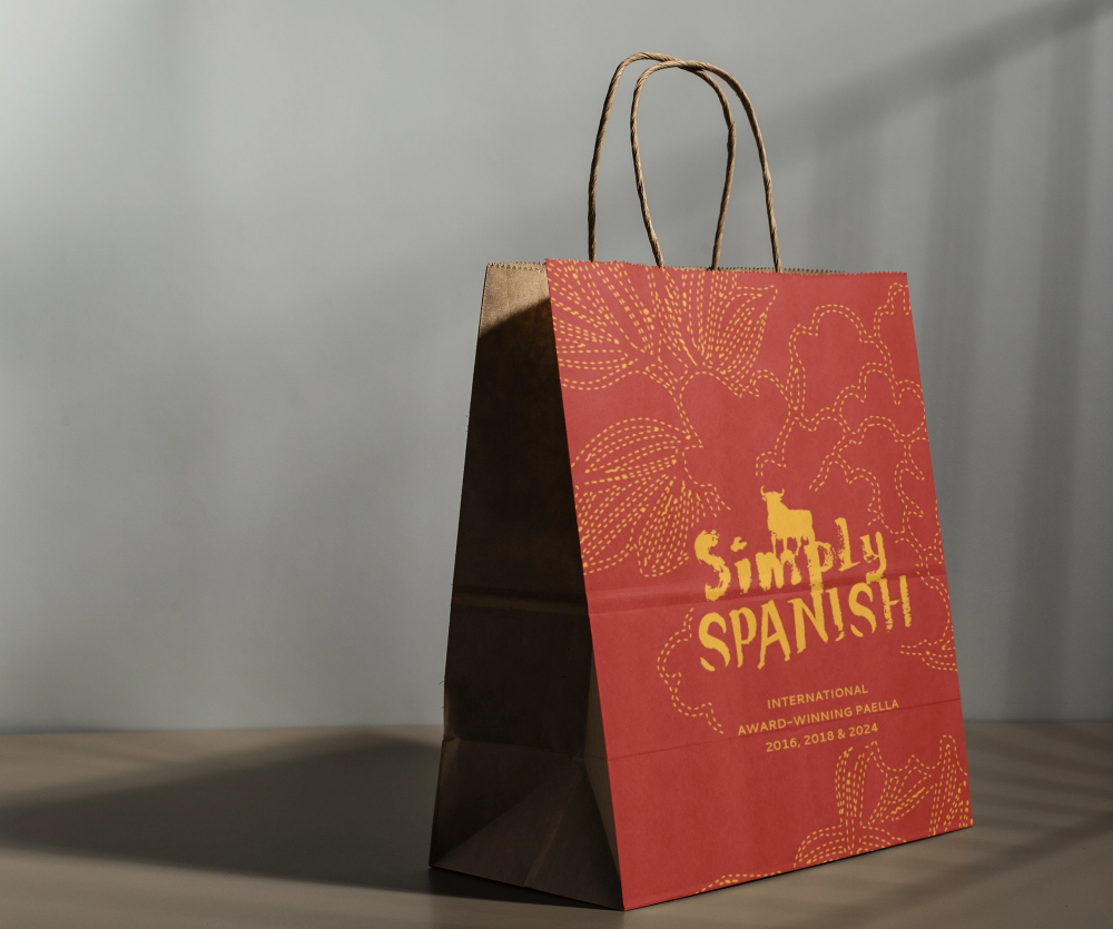 A paper shopping bag with red and brown colors. The red side features a graphic of leaves and the text 'Simply Spanish' along with details about paella awards.