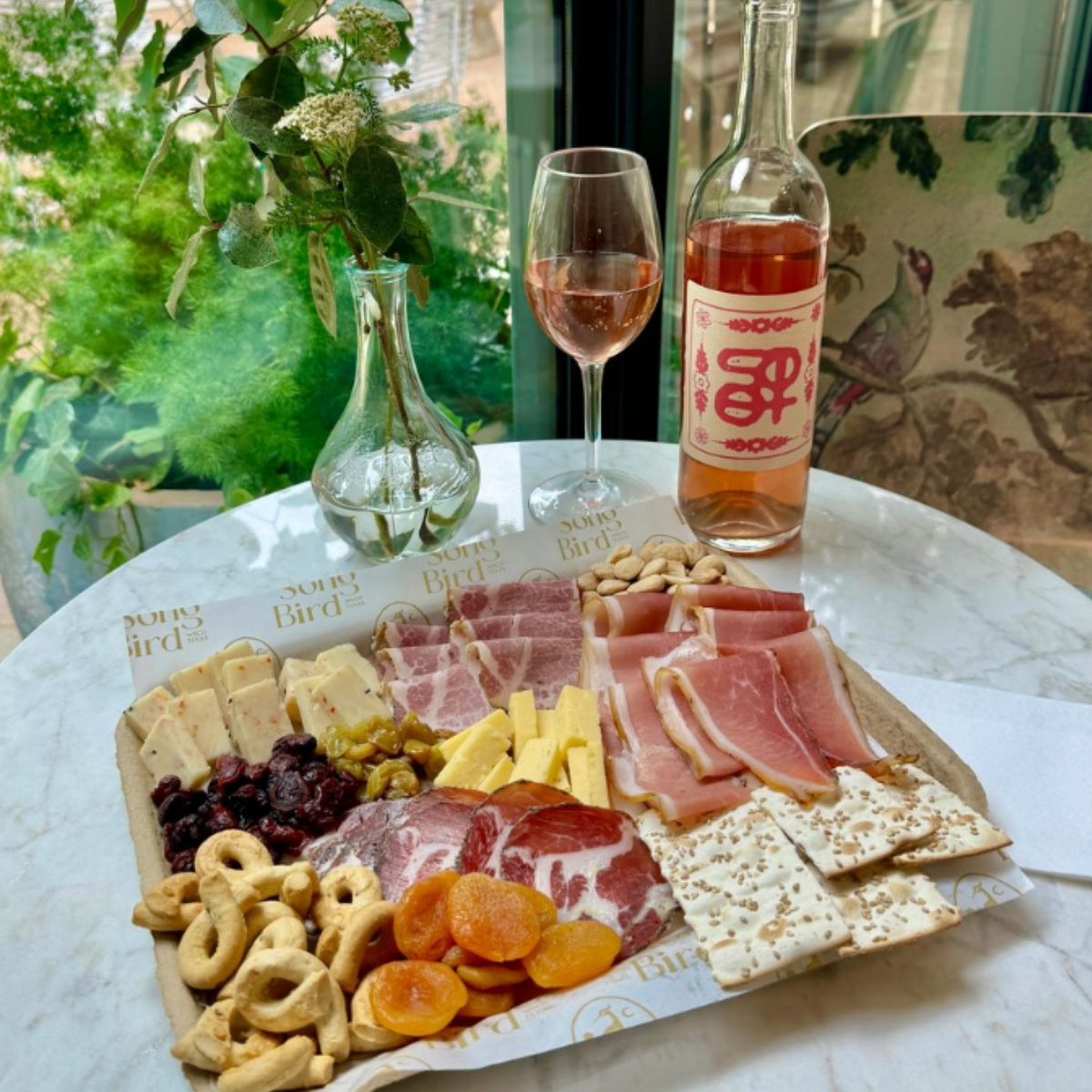 A charcuterie board with various meats, cheeses, dried fruits, and crackers, set on a marble table near a window with greenery outside. There is a glass of rosé wine, a bottle of rosé, and a vase with green and white foliage on the table.
