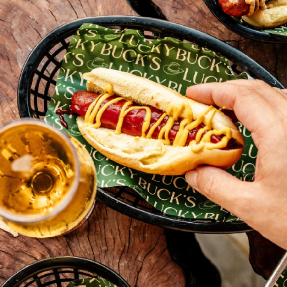 Hot dog with mustard in a bun on a green Lucky Buck's paper liner, hand holding the hot dog on a wooden table, with a glass of beer and other dishes visible.