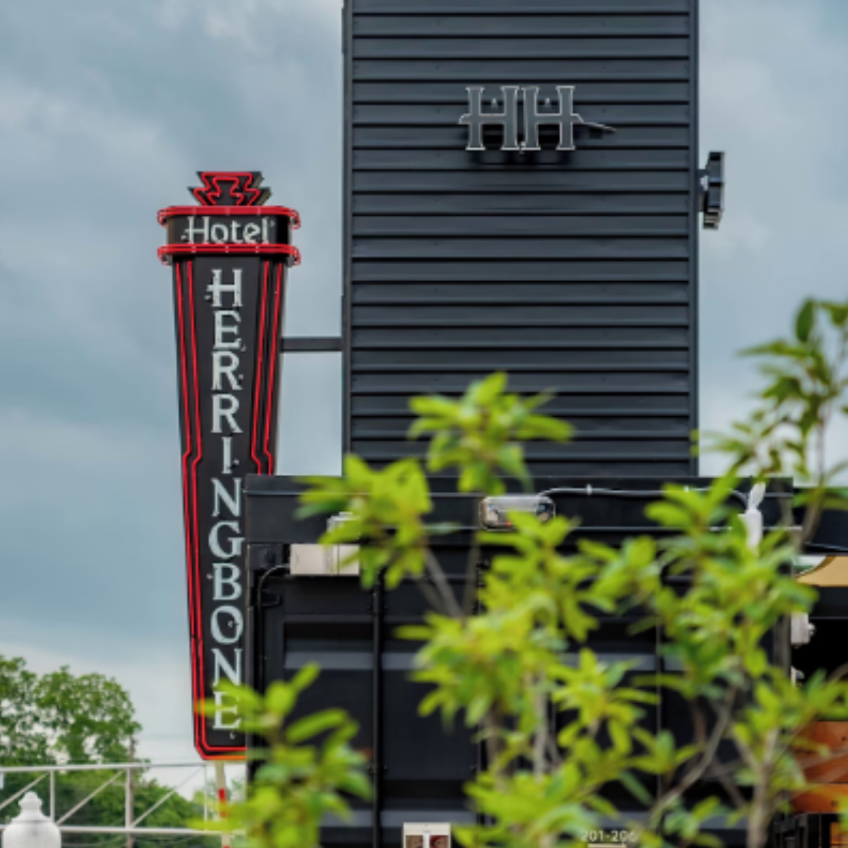 Hotel signage with a vertical neon sign reading 'HERRINGBONE' in white letters on a black background and the word 'Hotel' in smaller text at the top, set against a cloudy sky, with green foliage in the foreground.