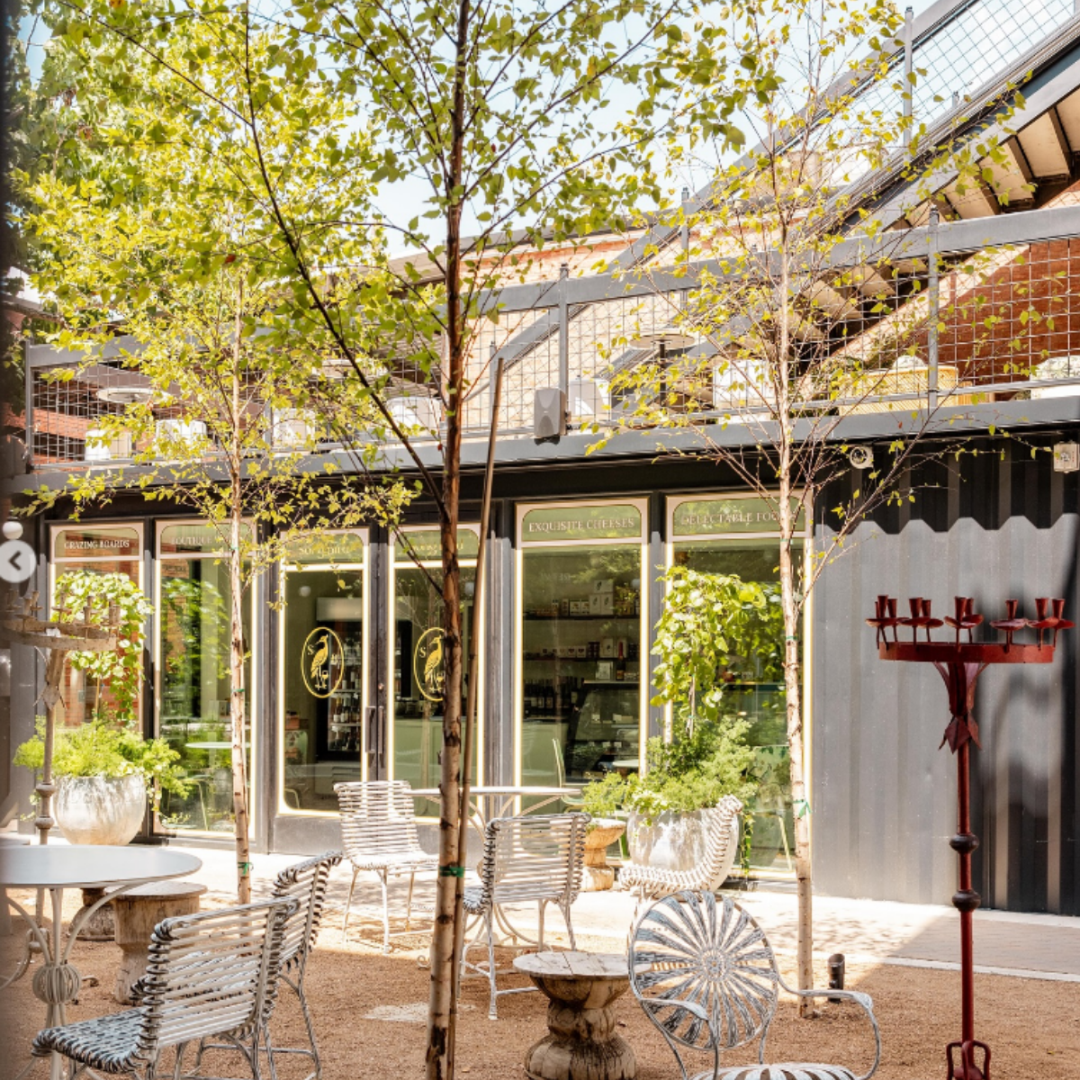 Outdoor seating area with chairs and tables in front of a storefront with glass doors, potted plants, trees, and a red coat rack, under trees and a building with a fire escape ladder.