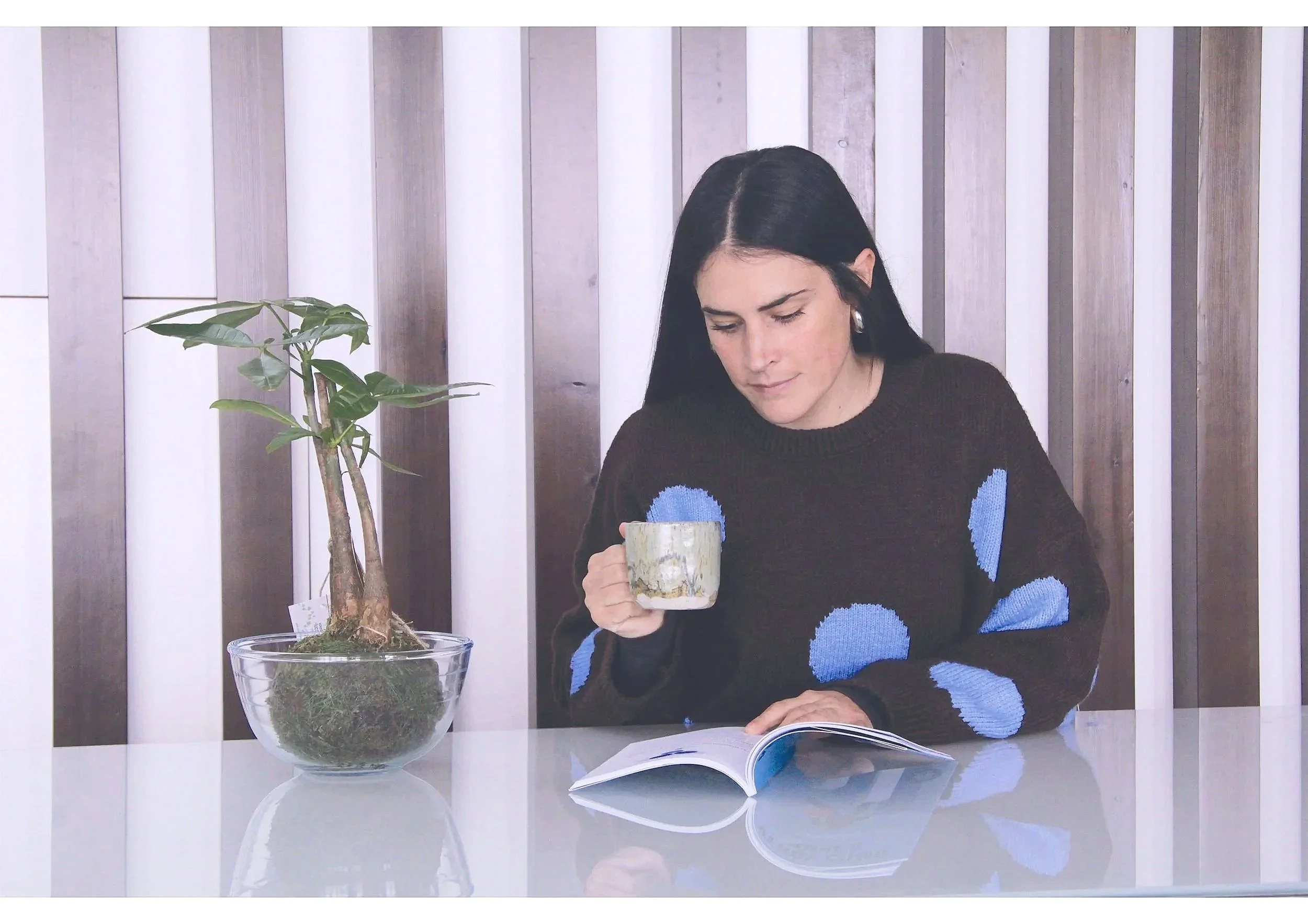 A woman with black hair, wearing a brown sweater with blue polka dots, sitting at a white table, holding a mug in her right hand, reading a magazine, with a potted bonsai tree on her left, against a wooden panel background.