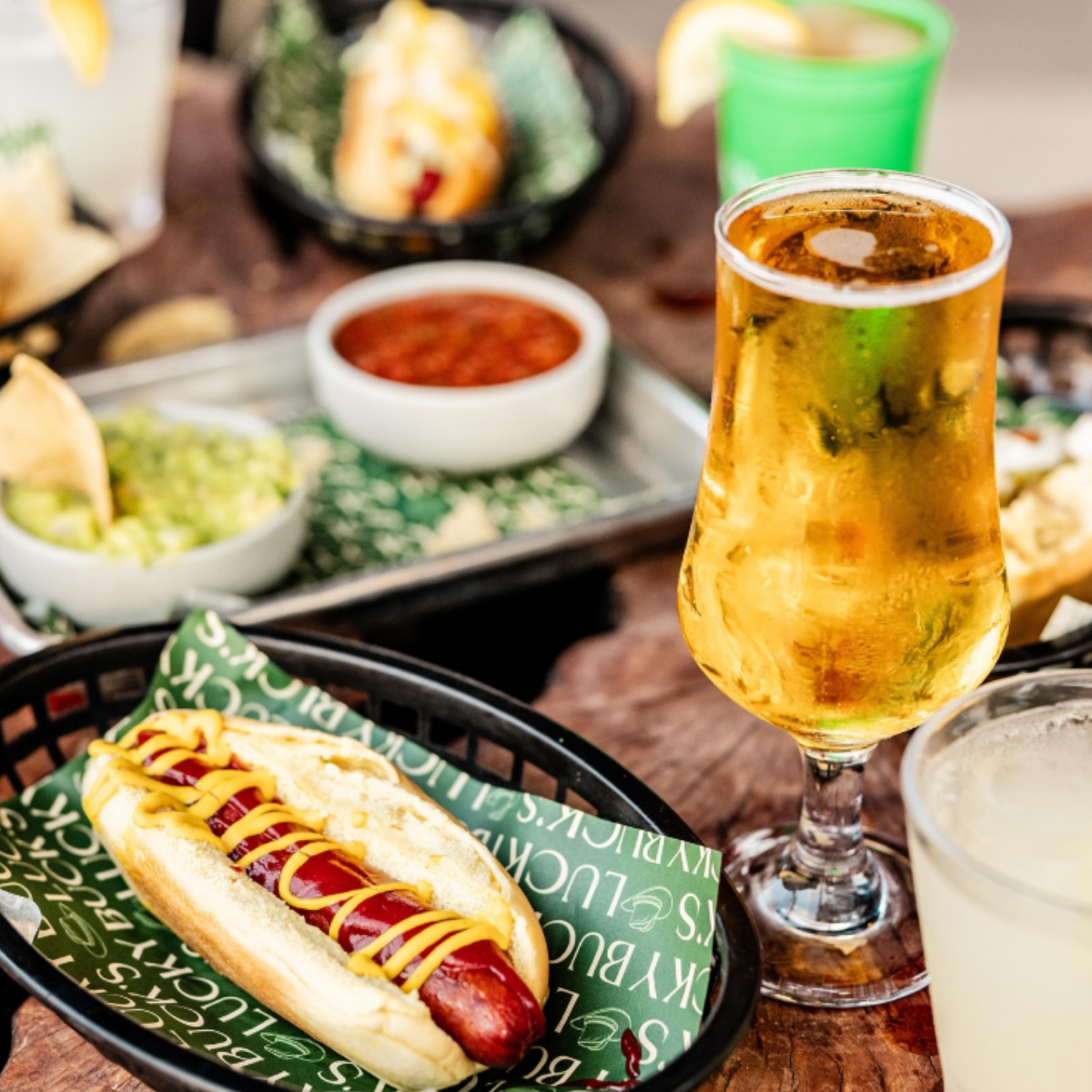 A hot dog with mustard in a bun on a tray, a glass of beer, a glass of a light-colored drink, salsa, guacamole, tortilla chips, and other condiments on a wooden table at a restaurant.