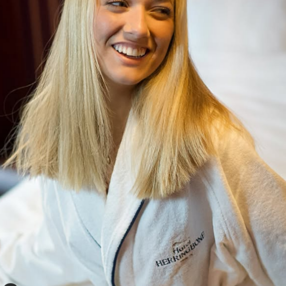 A smiling woman with long blonde hair wearing a white chef's jacket with the logo 'HERRINGBONE' embroidered on it.