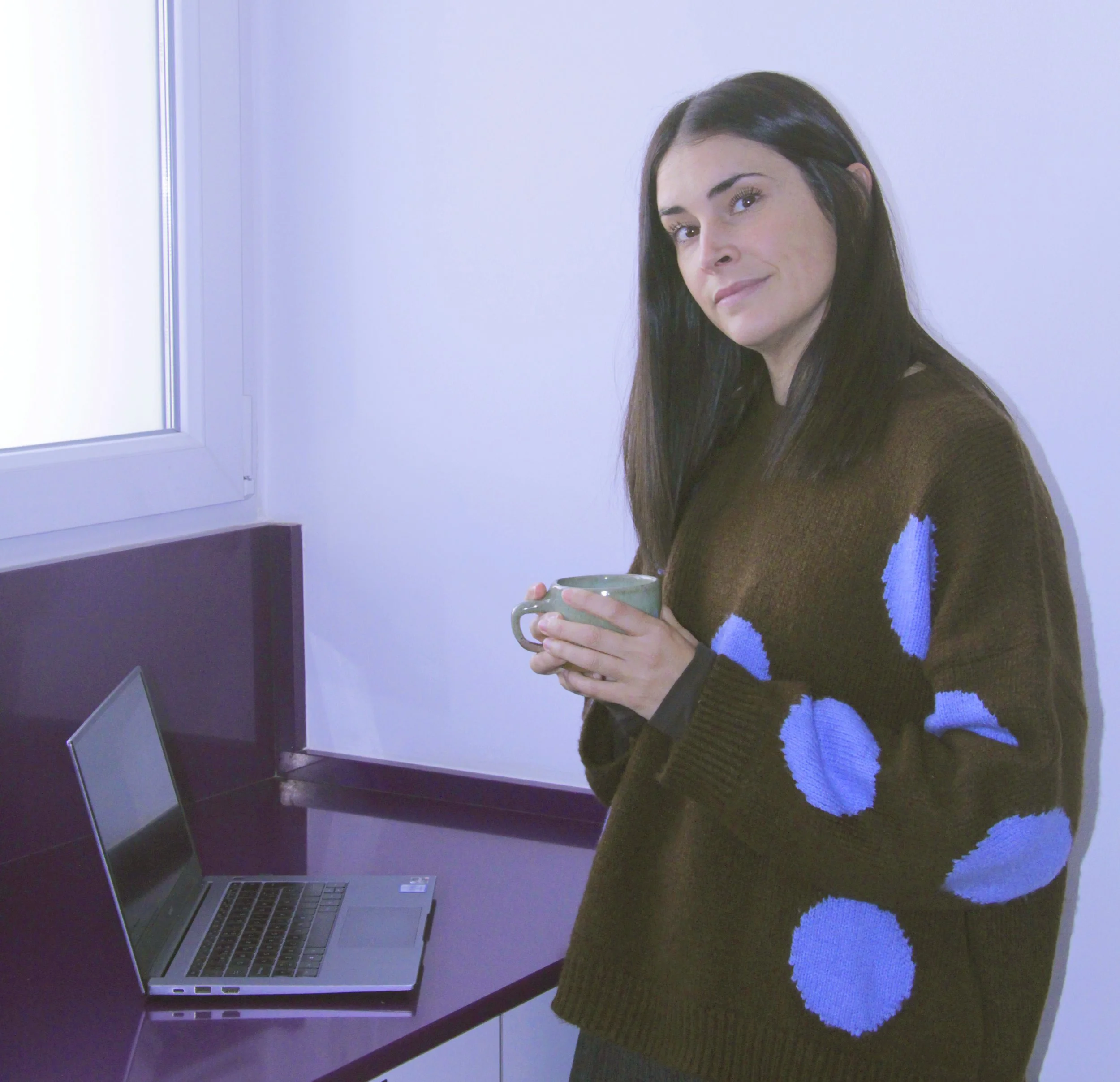 A woman with dark hair standing near a counter, holding a mug, with a laptop open on the counter.