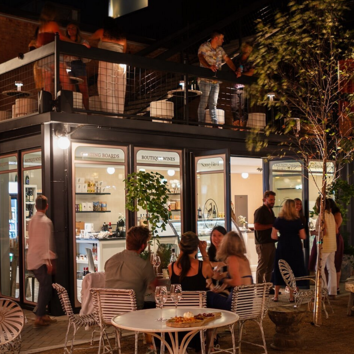 People socializing at an outdoor restaurant and bar at night, with some on the upper balcony and others seated at tables outside.