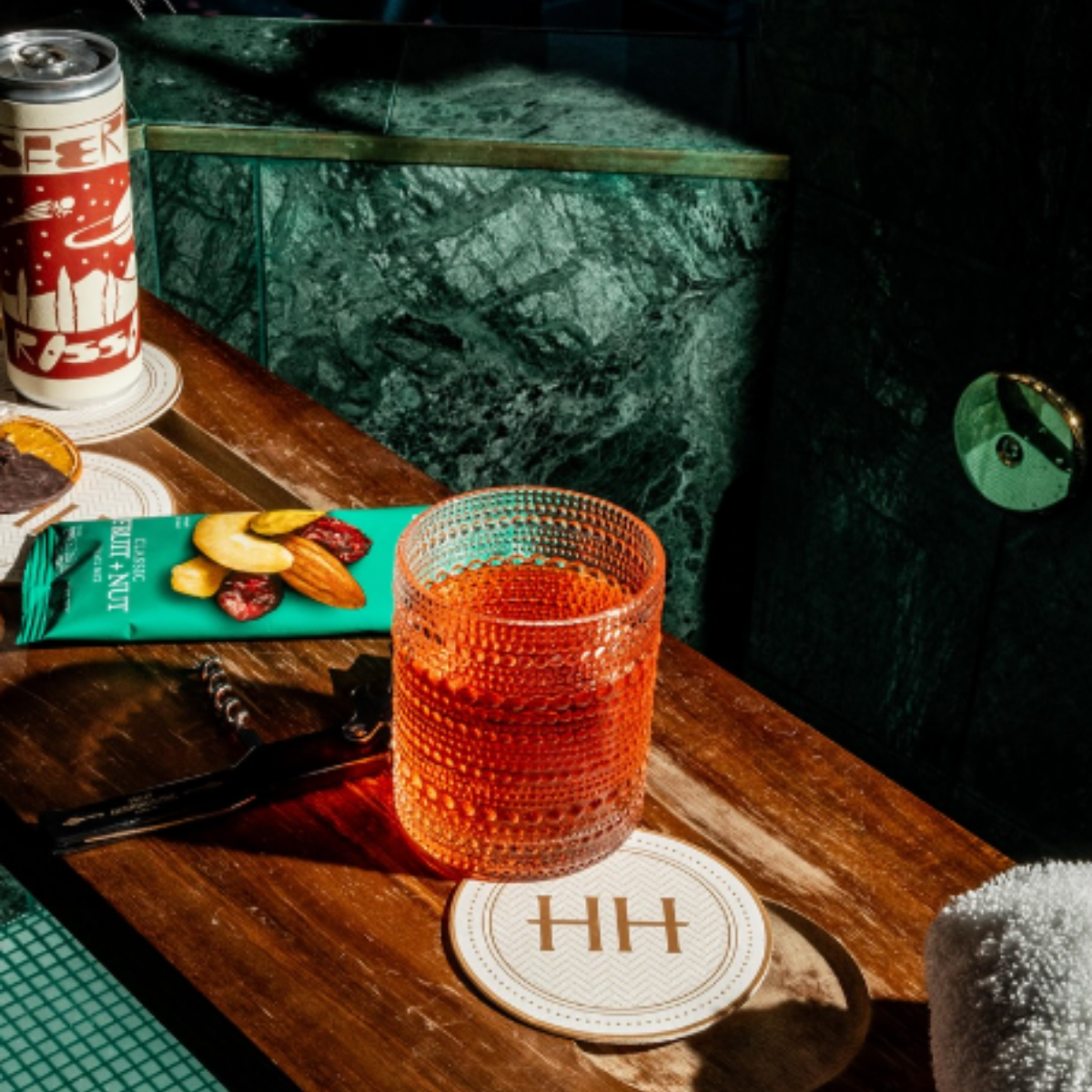 A wooden table with various items including an orange textured glass, a snack pack, a can, a small dish, and some utensils, with a marble wall and dark surroundings in the background.