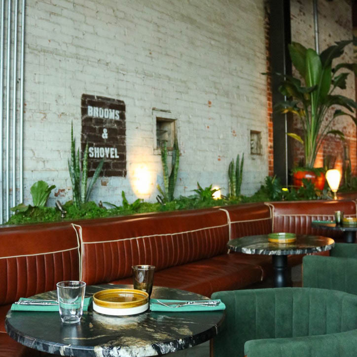 A cozy restaurant interior featuring a white brick wall with potted plants, a brown leather booth, and green upholstered chairs. There are tables set with plates, glasses, and cutlery, with warm lighting and a sign that reads 'Brooms & Shovel' on the
