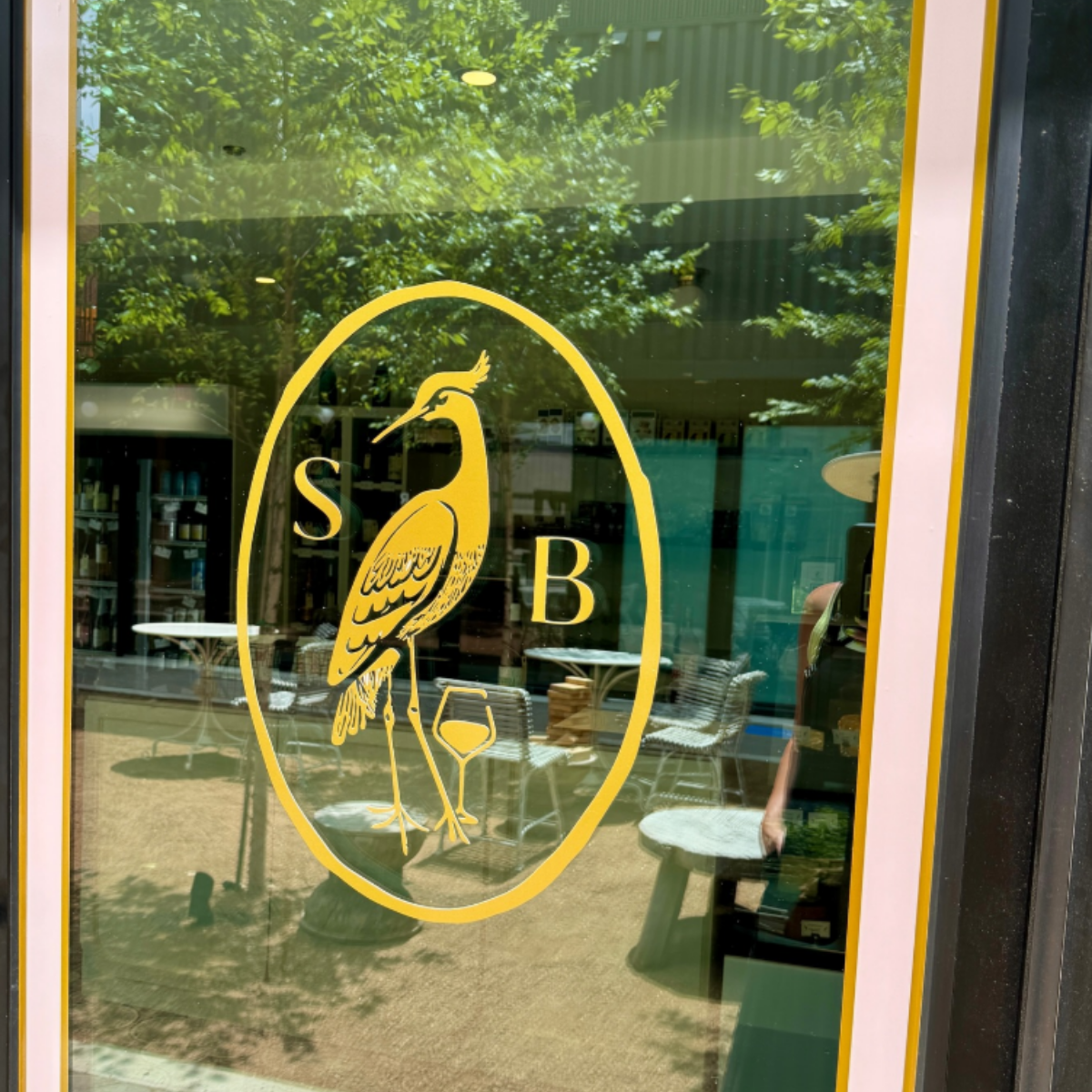 Glass door with yellow logo of a heron holding a glass, with the letters 'S' and 'B' on either side of the heron. The reflection of outdoor trees and furniture can be seen through the glass.