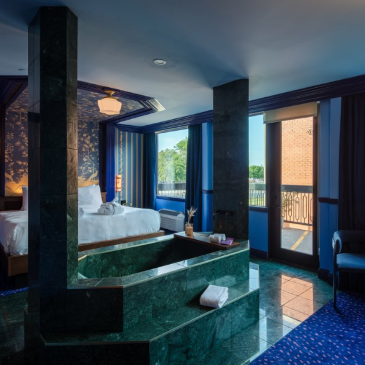 Hotel room with bed, green marble partition, large windows with blue curtains, and a balcony door showing outside view of trees and sky.