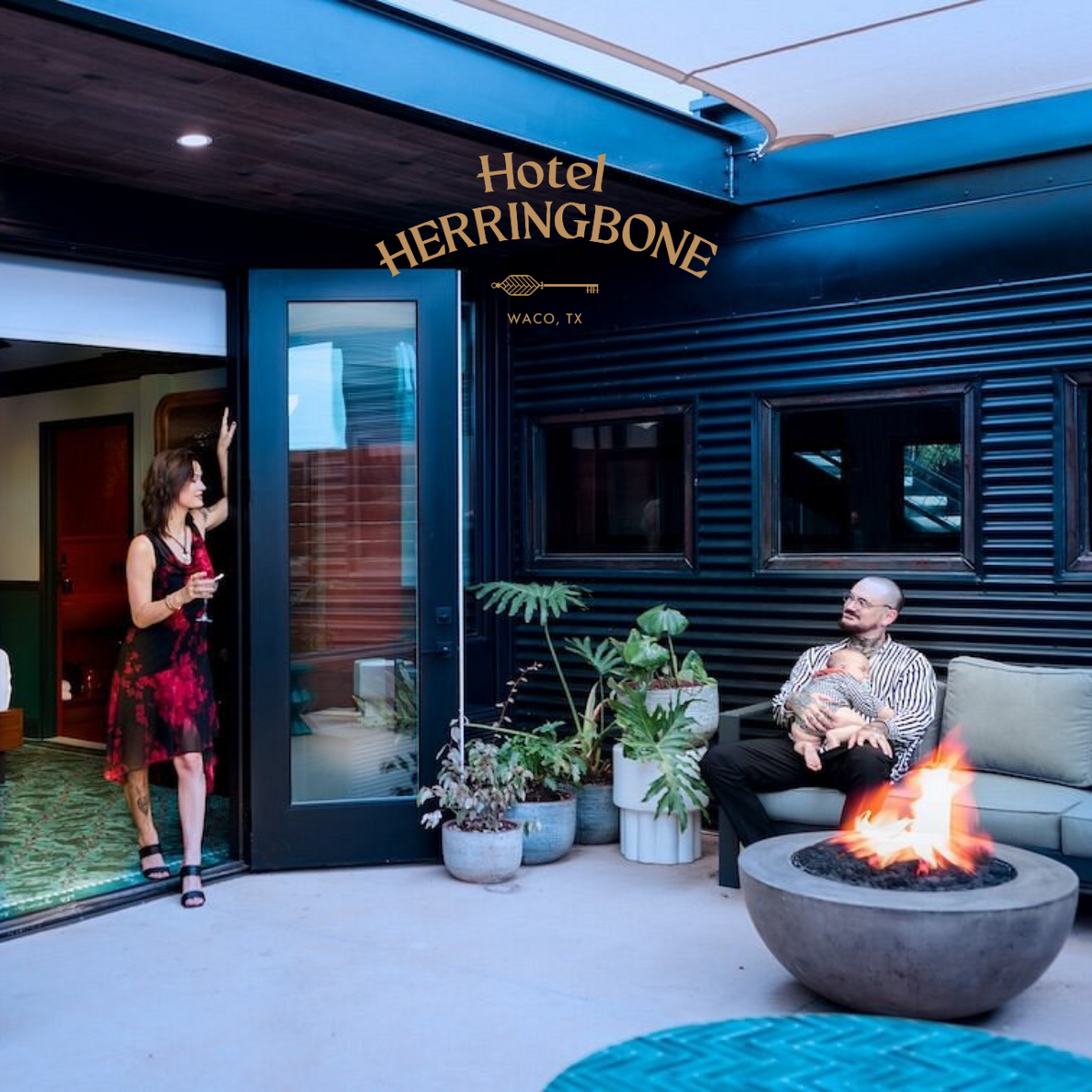A woman standing at the doorway of Hotel Herringbone in Waco, Texas, holding a glass of wine, while a man sitting on outdoor furniture holds a baby, with a fire pit and potted plants nearby.