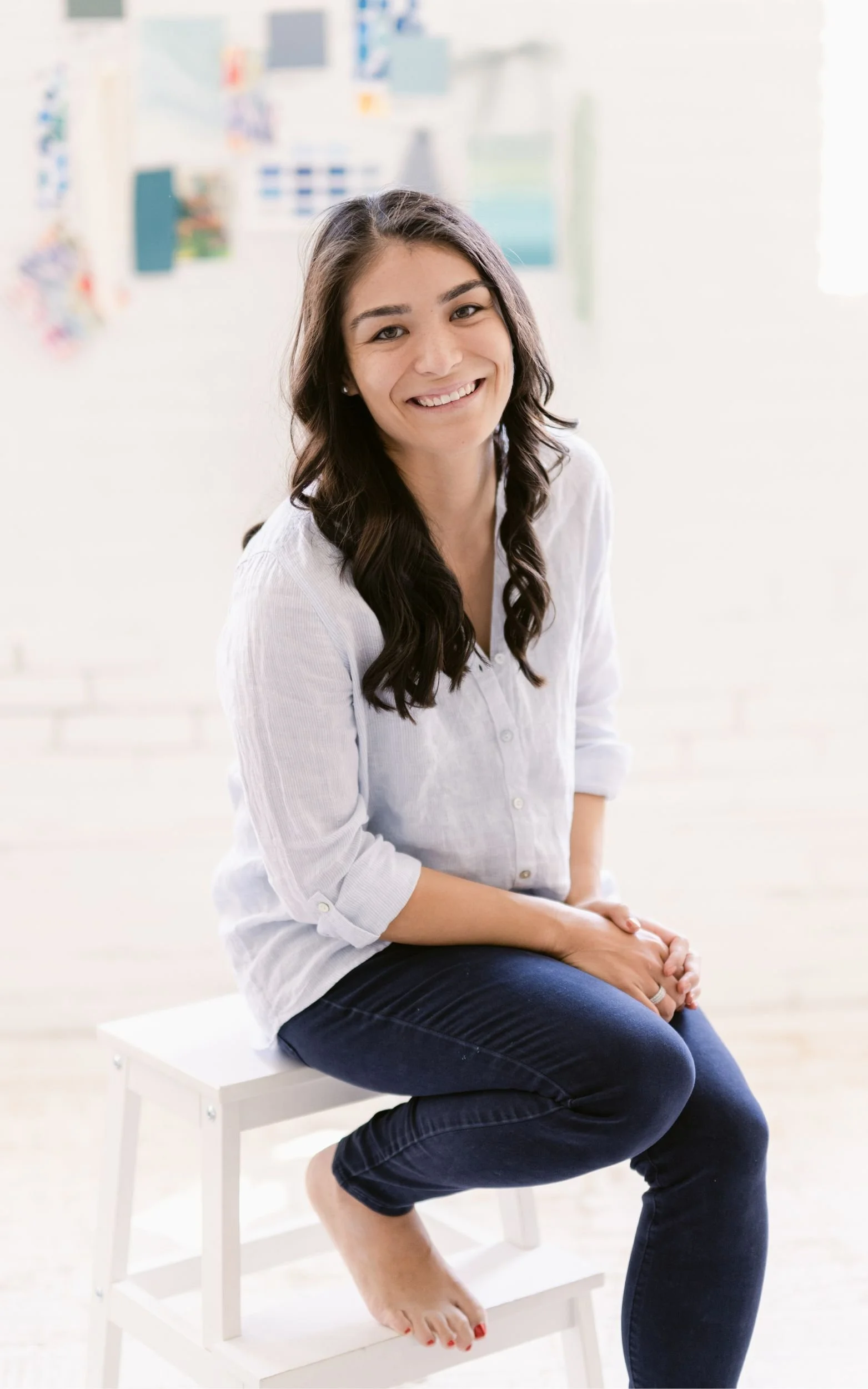 A smiling woman sitting barefoot on a white stool in a bright creative studio with inspiration swatches on the wall behind her.