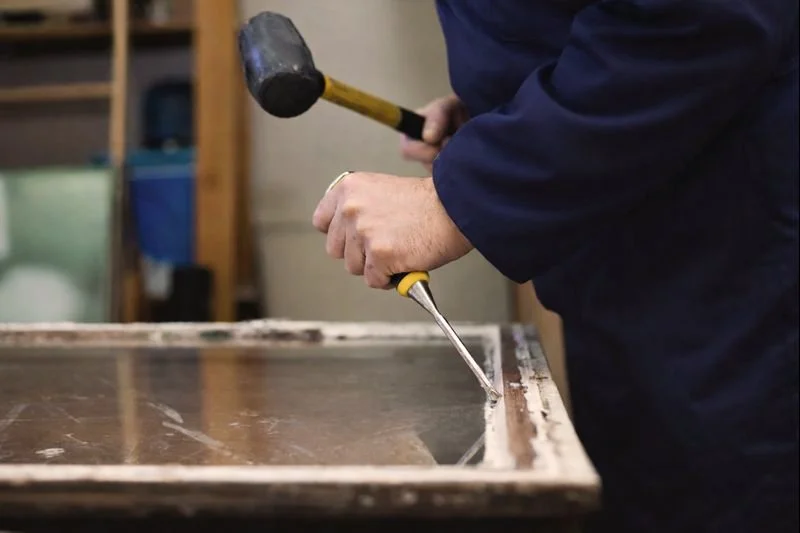 Person repairs a window with a hammer and chisel in a workshop.