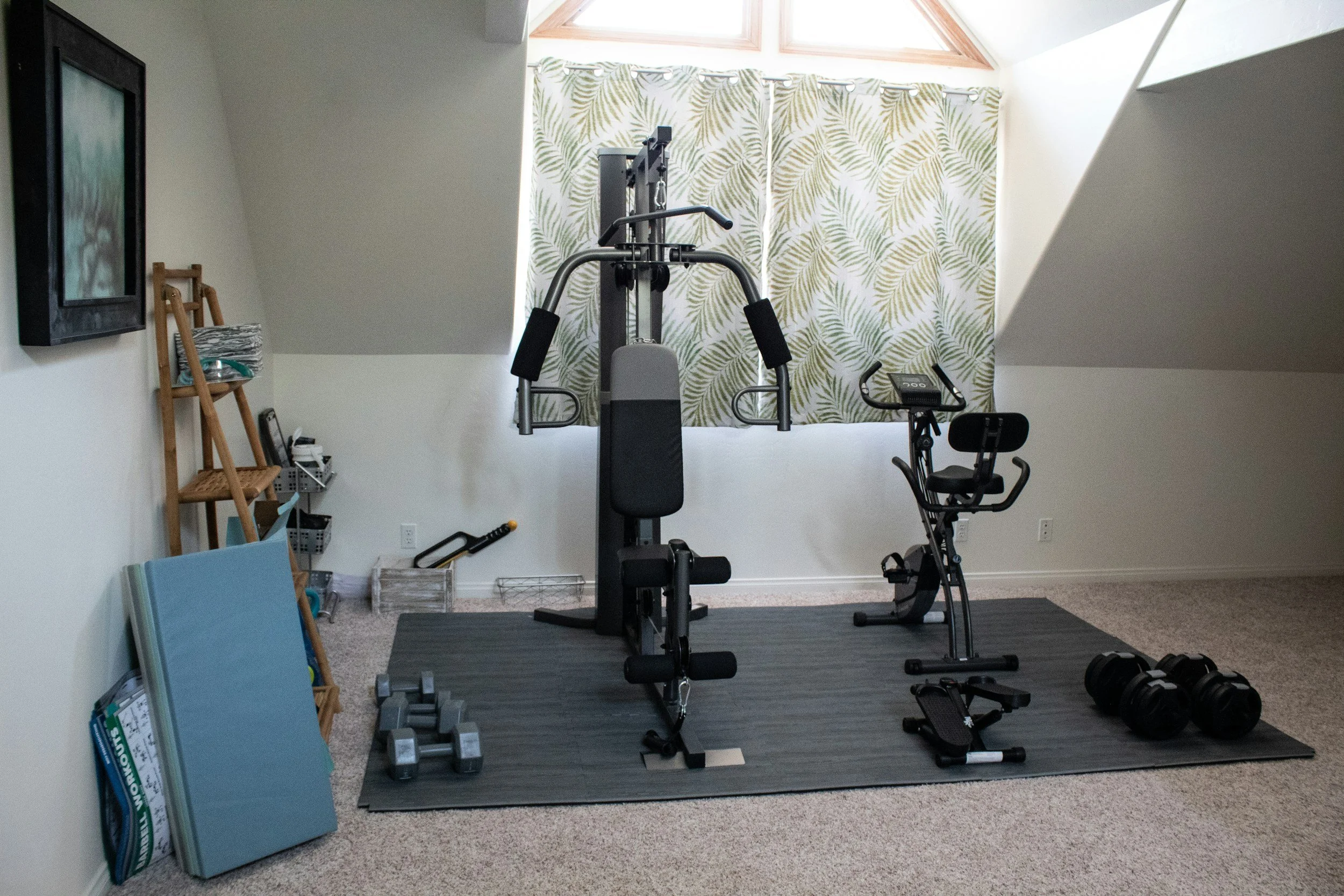 Home gym with a multi-function workout machine, exercise bike, dumbbells, and yoga mats on a black mat, next to a wall with a window and curtain featuring green leaf patterns.