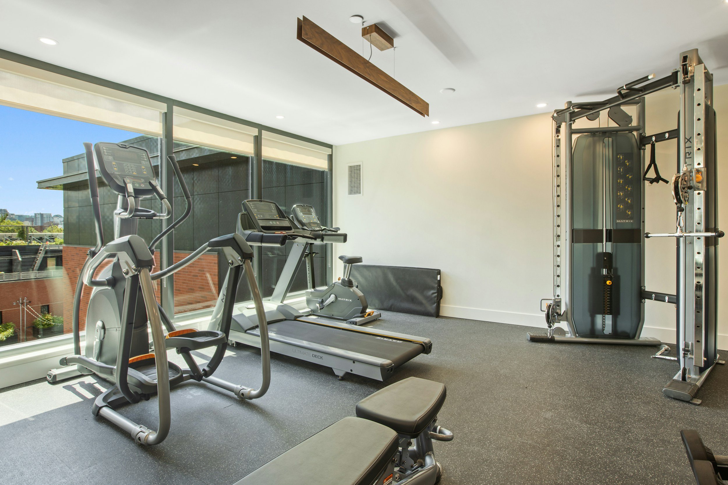 Modern indoor gym with cardio equipment including a treadmill, stationary bike, and elliptical machine near large windows, with a wall-mounted piece of exercise equipment on the right.