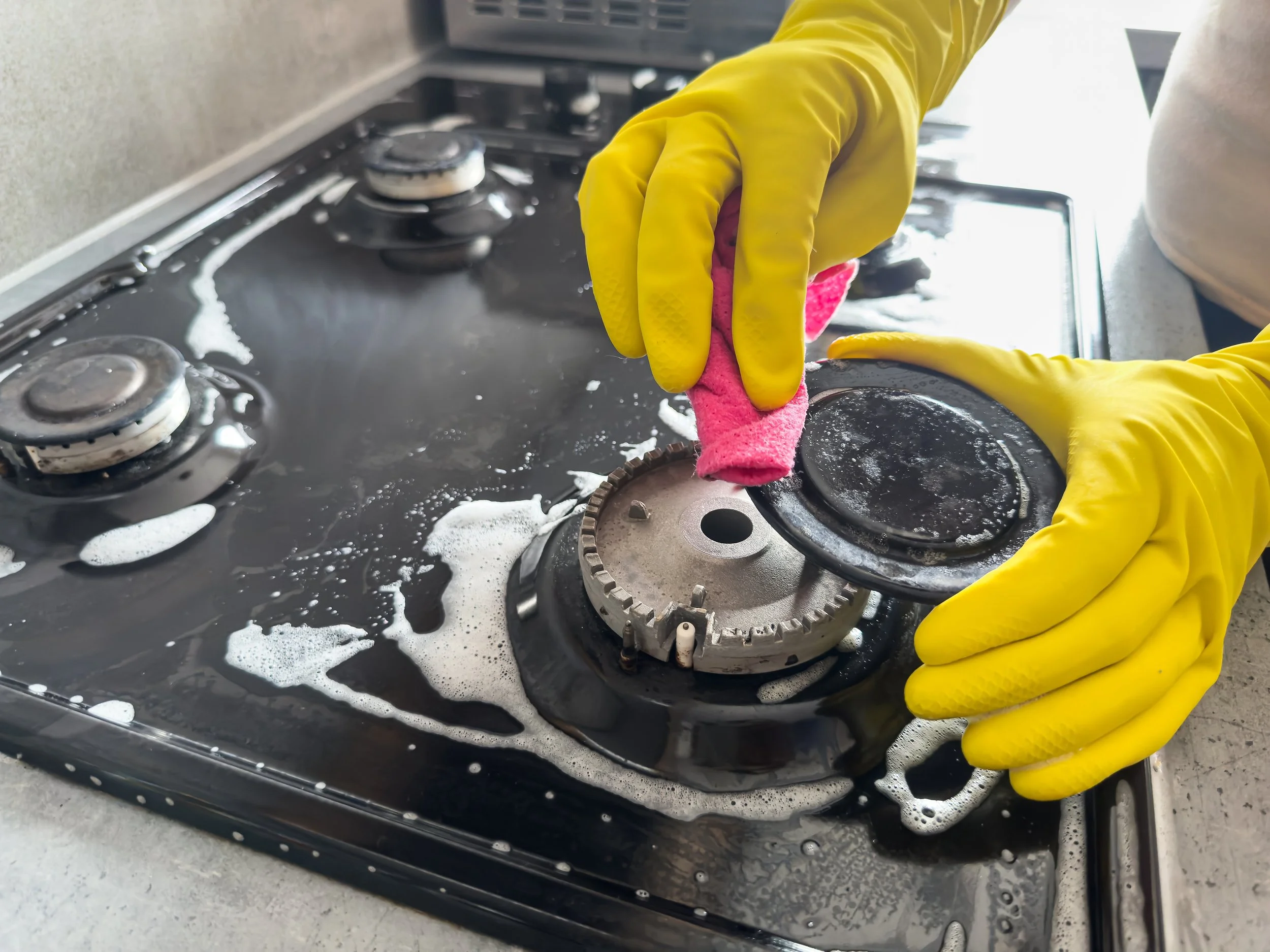 Person wearing yellow rubber gloves cleaning a black stovetop with a pink cleaning sponge and soapy water.