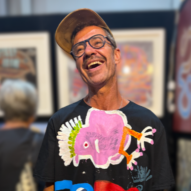 A smiling man wearing glasses, a baseball cap, and an artistic Aboriginal-designed T-shirt with a pink Australian bird design at an art gallery.