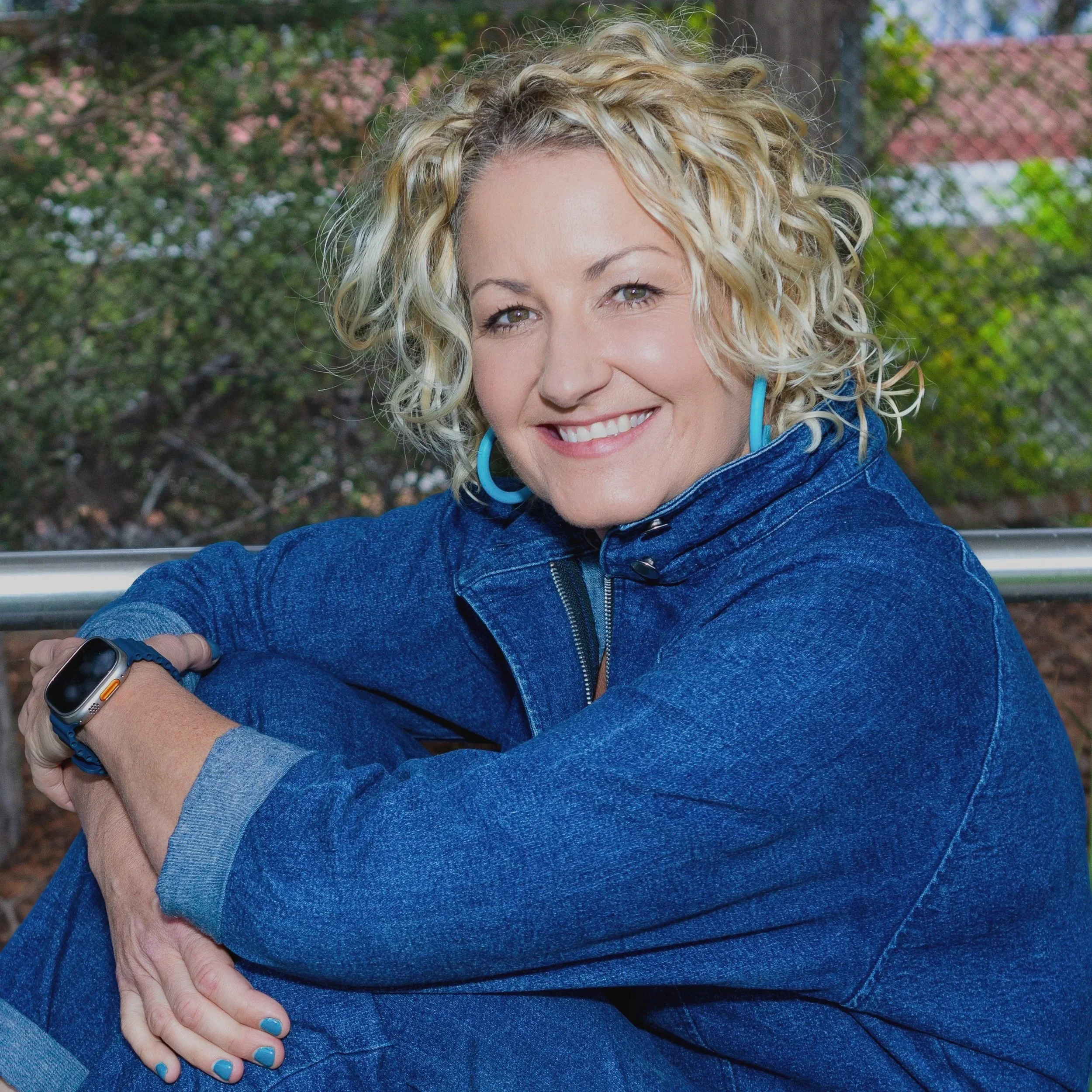 Smiling woman with curly blonde hair wearing a blue denim jacket and blue earrings, sitting outdoors with trees and a fence in the background.