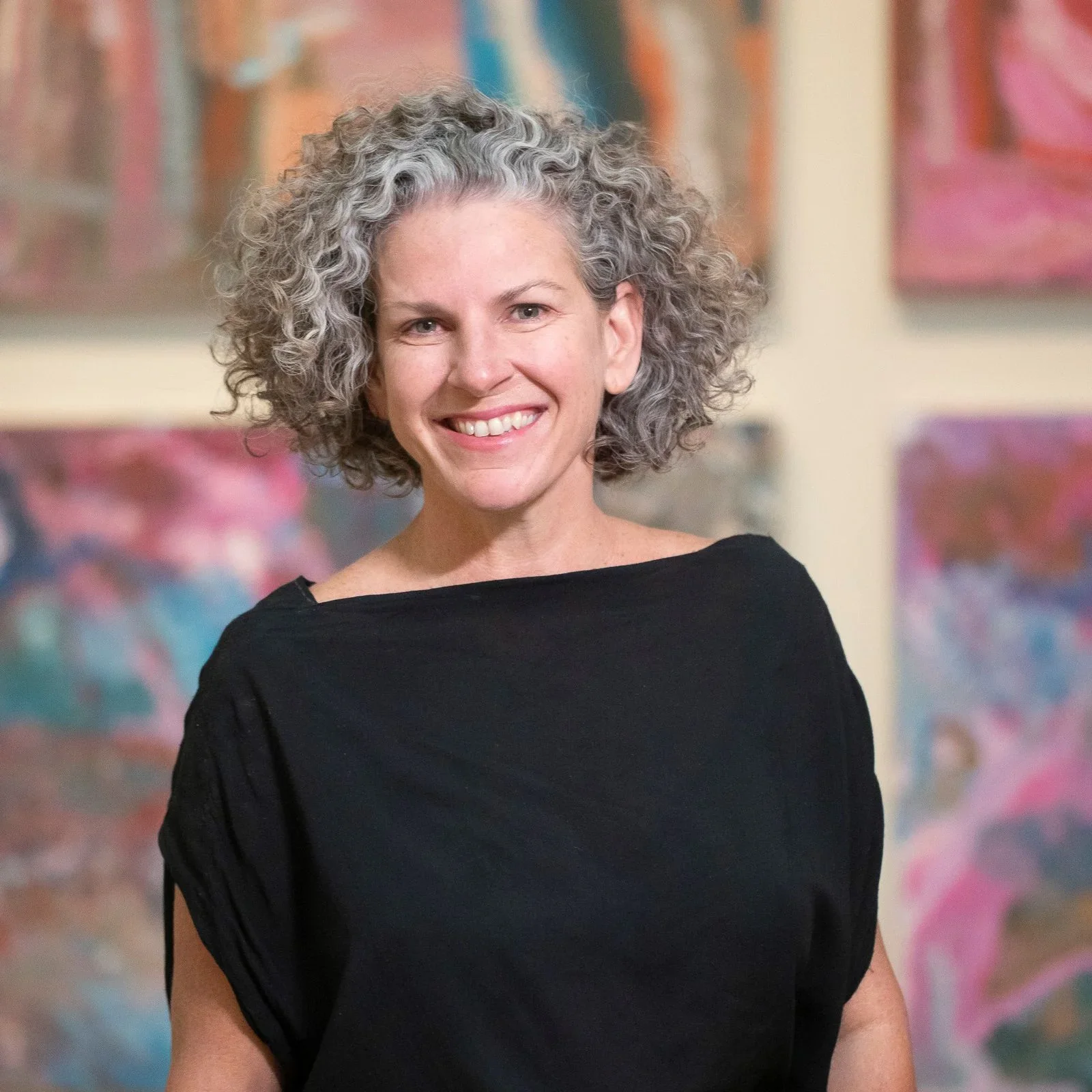 A woman with curly gray hair smiling, wearing a black top, standing in front of a colorful abstract painting.