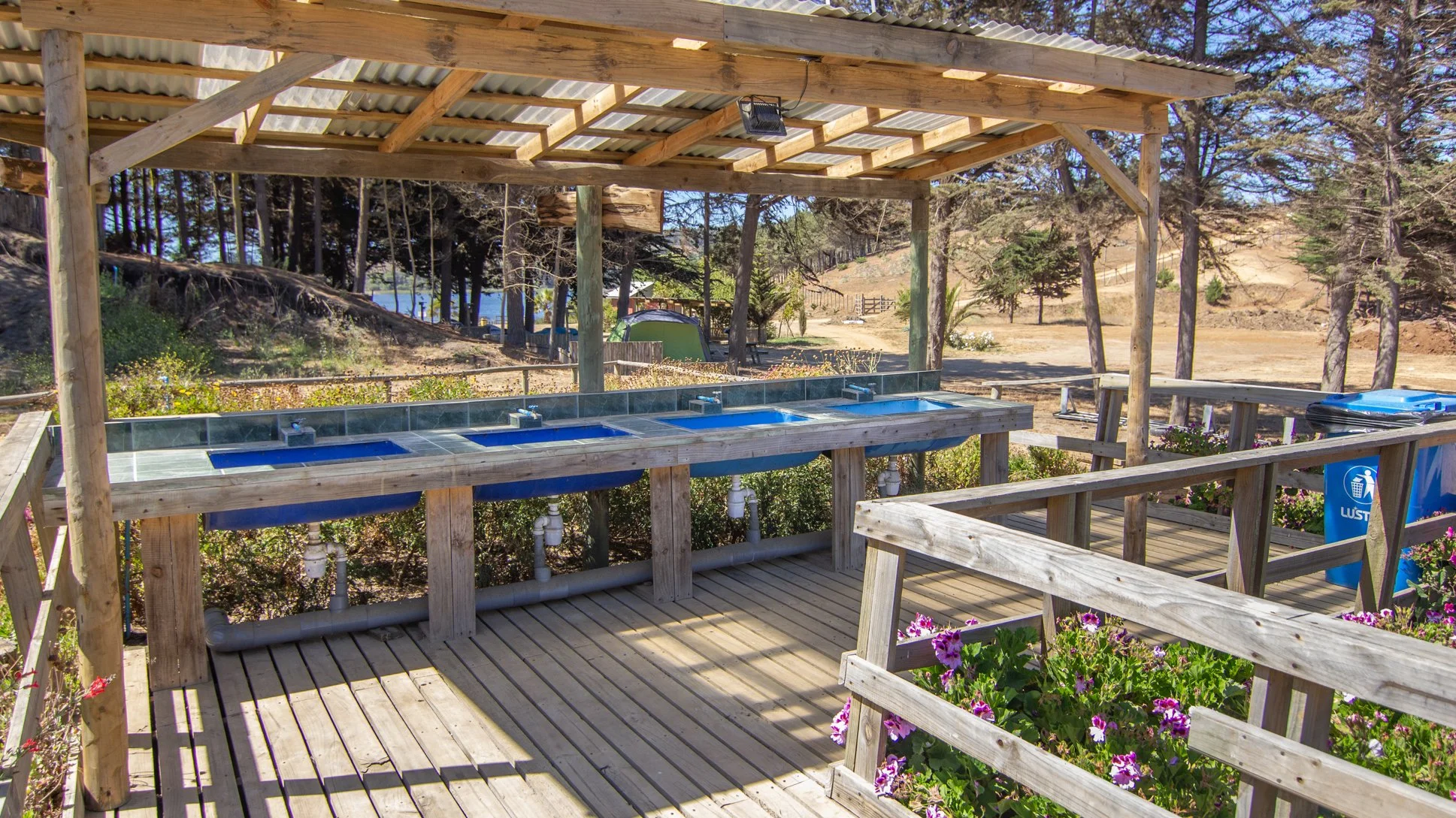 Lavaderos públicos en un área al aire libre con estructura de madera, con varias lavadoras azules y una papelera azul con símbolo de basura.
