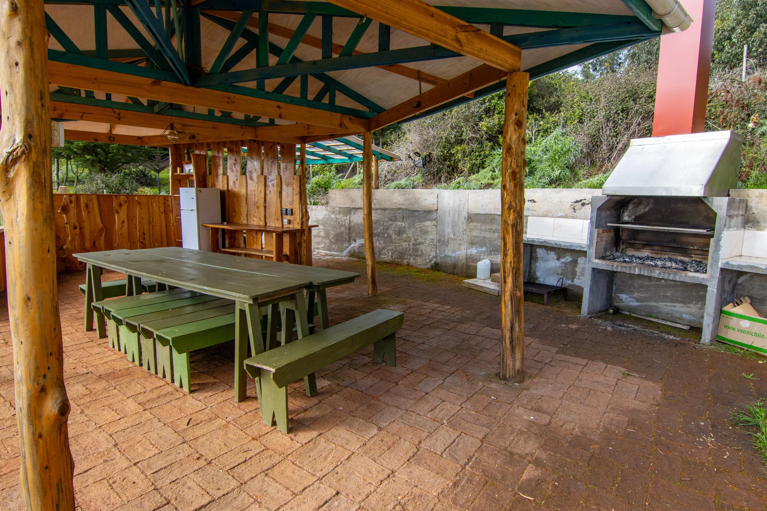 Una zona de asador al aire libre con una parrilla de carbón, mesa de madera con bancos y una estructura de madera con techo, rodeada de naturaleza.